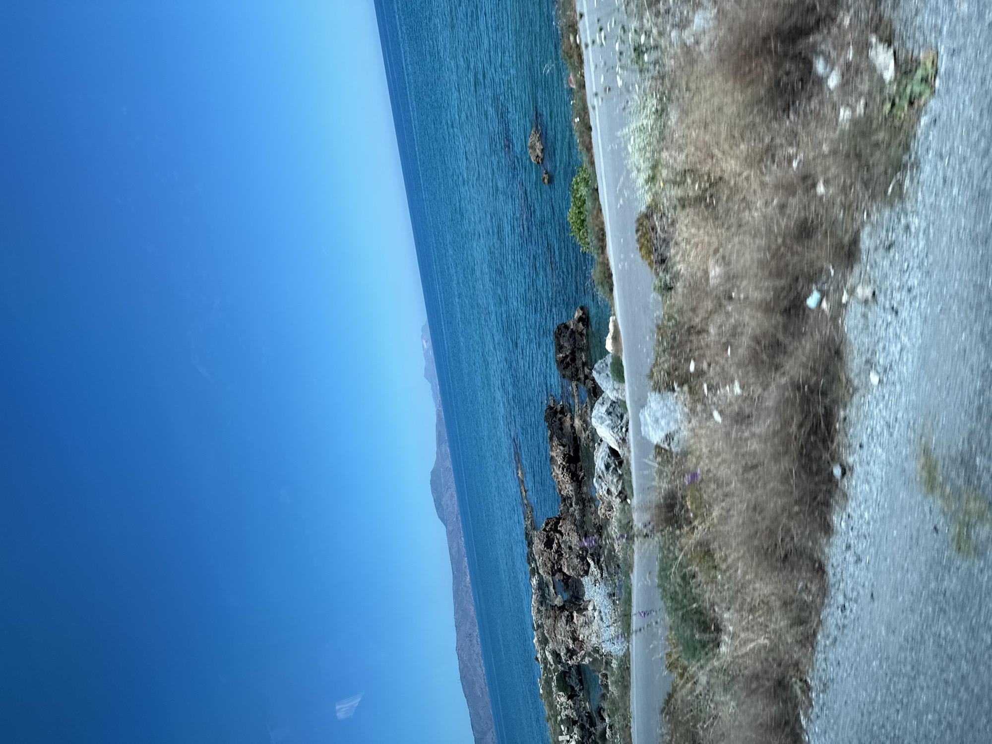 The image shows a coastal road in Rethymno, Greece, on a clear, sunny day. The sky is a vibrant, cloudless blue. The sea is a deep turquoise, with gentle waves breaking against a rocky shoreline. Mountains are visible in the distance, forming a hazy outline against the horizon. The road itself is paved and appears to be winding along the coast. On the side of the road, there is dry, scrubby vegetation, with some scattered rocks and wildflowers. The scene suggests a natural, undeveloped coastal area, likely a popular spot for scenic drives or exploring the natural beauty of the region.