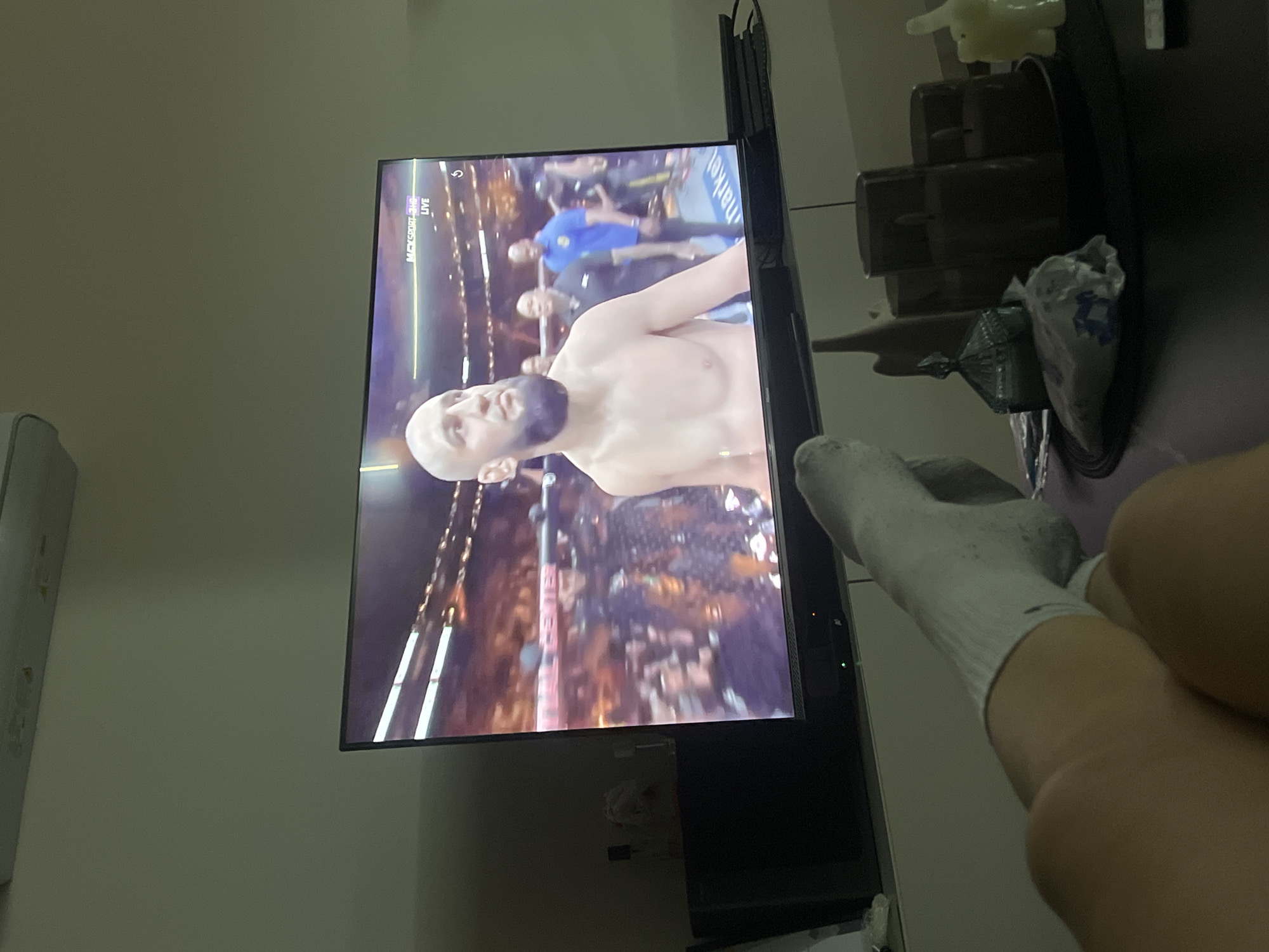 A television screen displays a live broadcast of a mixed martial arts event. The featured fighter is shirtless, with a shaved head and a beard, standing in a fighting ring before a crowd. A person's leg and foot, wearing a white sock, are positioned in front of the television. Next to the television is a small table with various items, including what appears to be a small toy and a decorative container. The scene is indoors, in a residential setting, although there is no specific visual information to associate it with Plovdiv, Bulgaria.