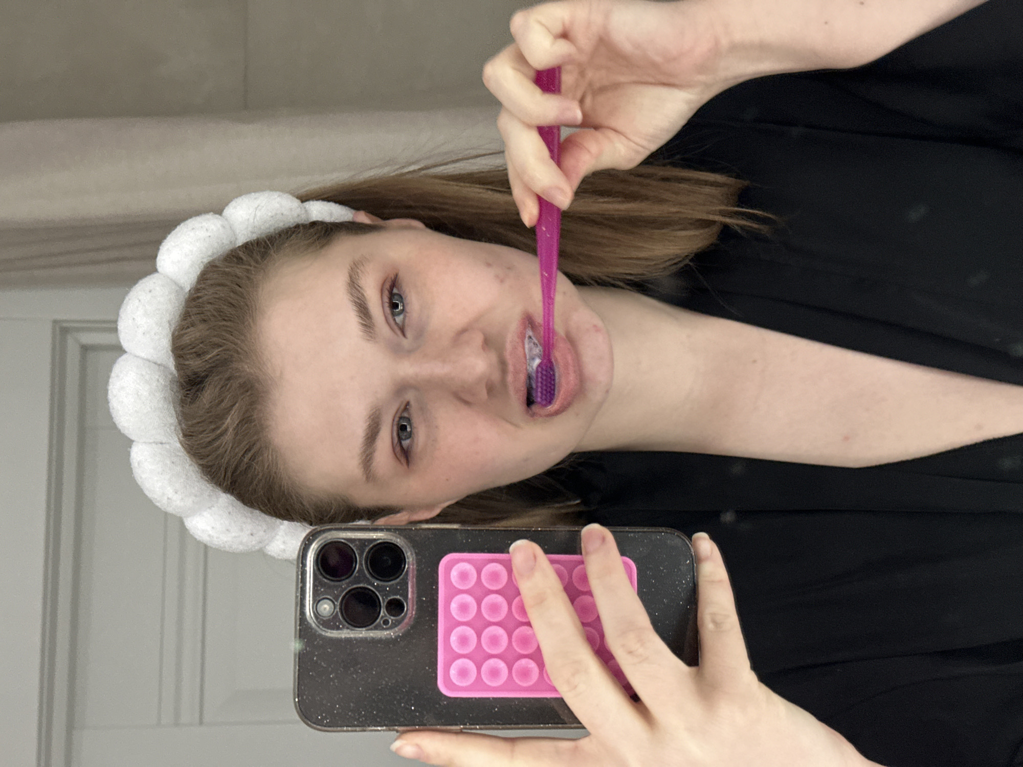 A young adult female is depicted in a mirror selfie taken indoors in Kazan, Russia. She has light brown hair, secured with a white, puffy headband, and is wearing a dark, possibly black, robe or garment. Her skin shows visible blemishes on her cheeks and chin. With her left hand, she holds a dark-colored smartphone, featuring a glittery case and a bright pink pop-it style accessory, angled to capture her reflection. Simultaneously, she is brushing her teeth with a purple toothbrush held in her right hand, with white toothpaste foam visible around the brush head and her partially open mouth. The background shows a light-colored wall with white architectural molding on the left and a light-colored, possibly textured wall or curtain on the right.