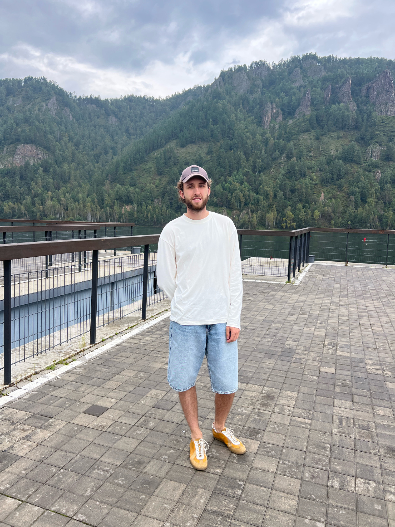 A man wearing a white long-sleeved shirt, denim shorts, a baseball cap, and yellow sneakers stands on a paved walkway. He is positioned in the foreground, facing the camera with his hands behind his back. Behind him, a body of water is visible, bordered by a dark railing and a pier with multiple levels. In the background, a dense, green forest covers a steep mountainside. The sky above is overcast. The scene is likely the embankment area of the Krasnoyarskoye Reservoir in Divnogorsk, Russia, near the Divnogorsk Hydroelectric Power Station.
