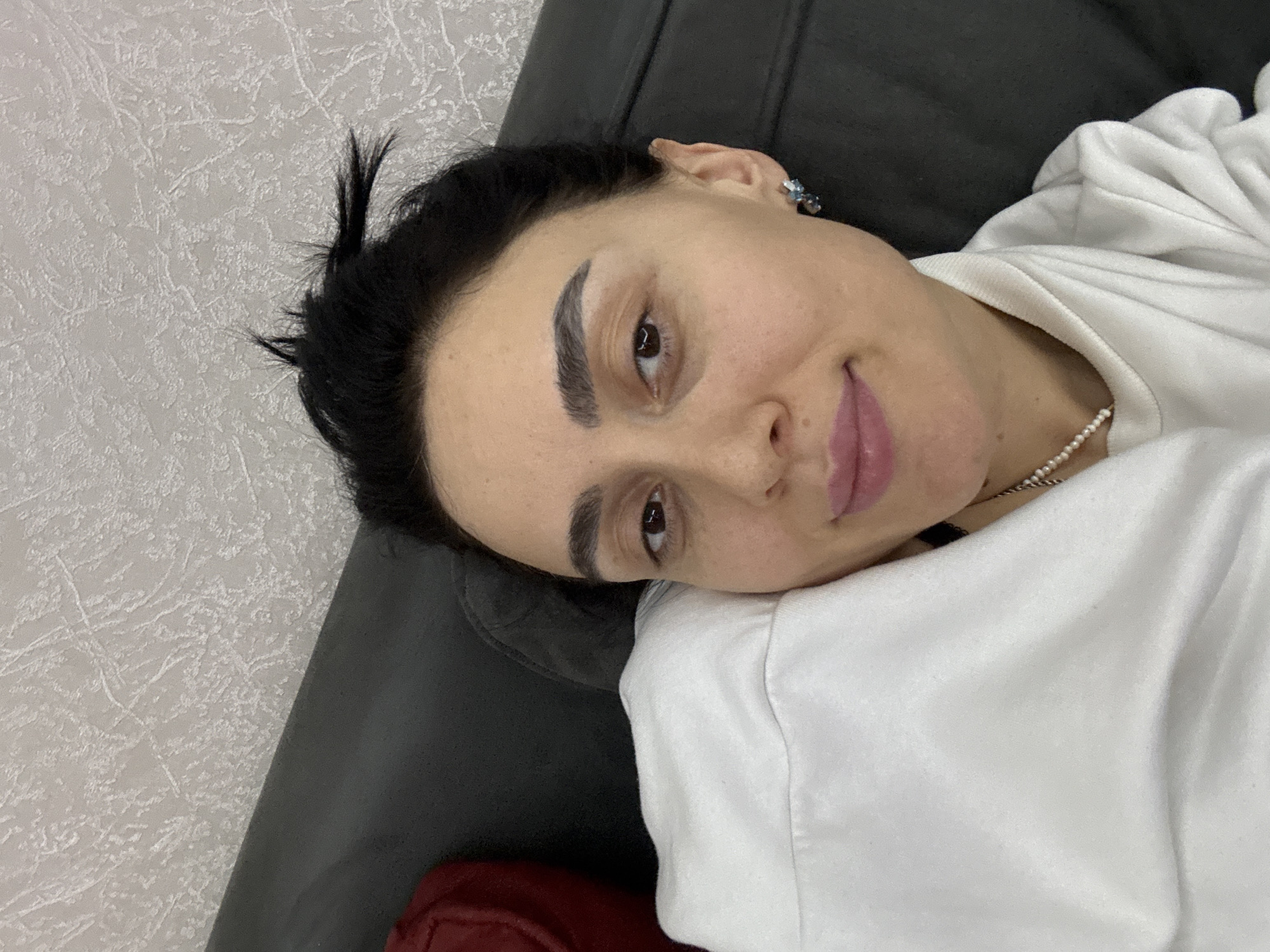 A medium shot shows a person lying on a gray sofa with a white wall in the background. The person has dark hair, dark eyebrows, and is wearing a white top with a pearl necklace. The person is looking at the camera with a slight smile. An earring is visible in their ear. A red object is visible in the bottom left corner of the frame. The location is unknown.