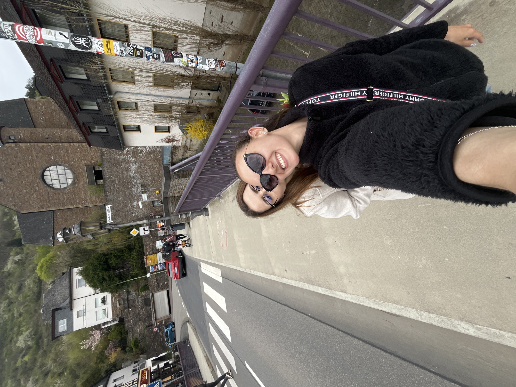 Two women are taking a selfie on a bridge in Cochem, Germany. The bridge has a purple railing. A street runs below with a crosswalk, a red car and a blue car are visible. Buildings line the street, including a stone building with a round window. A utility pole covered with stickers stands near the railing. The women are wearing sunglasses. One woman has a Tommy Hilfiger strap on her jacket.