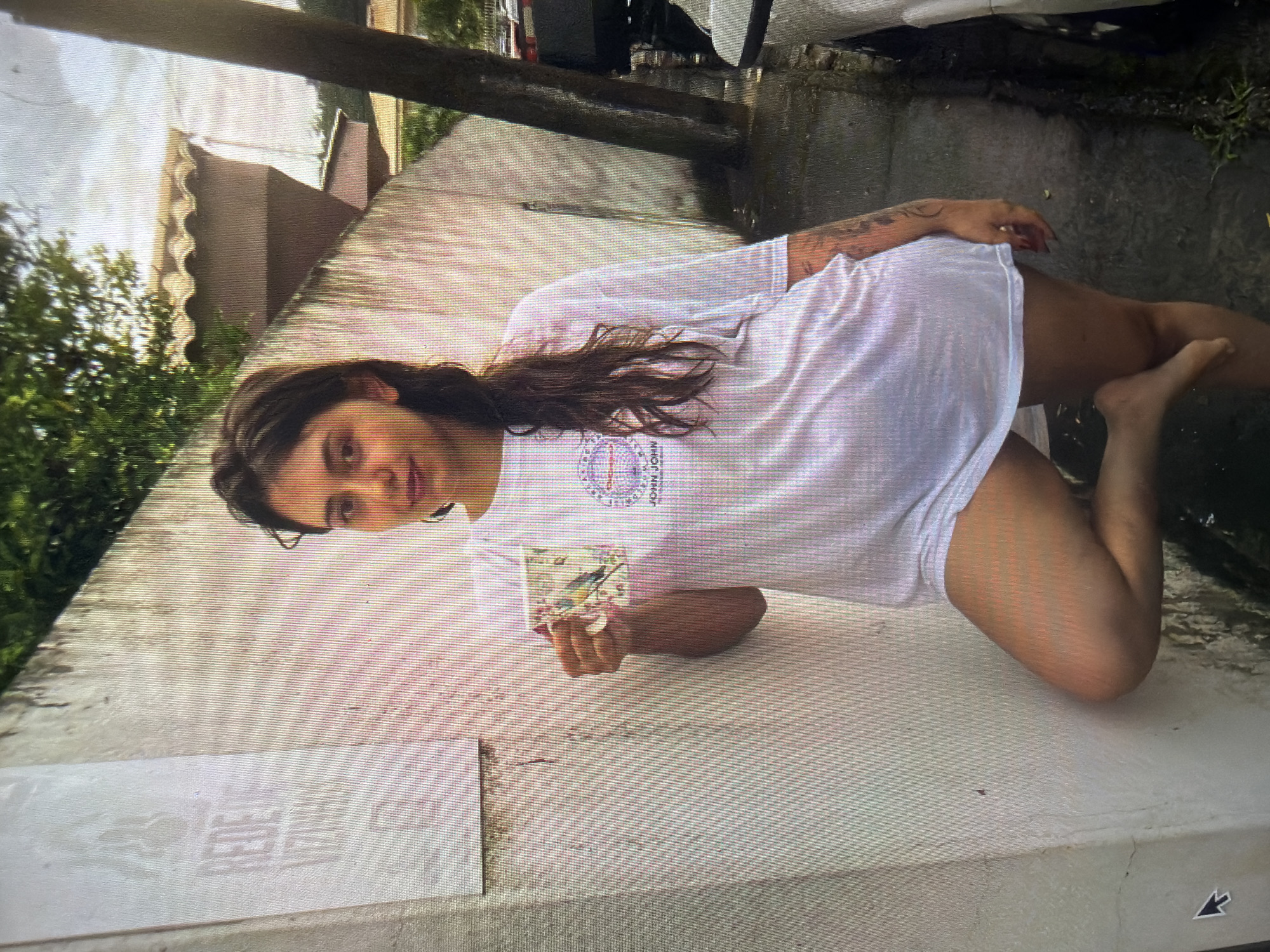 A young adult female is depicted standing, leaning her left side against a light-colored wall outdoors. She possesses dark hair, styled down, and is barefoot. Her attire consists of an oversized, loose white t-shirt that extends to her mid-thighs. A tattoo is visible on her left forearm. She holds a white mug adorned with a colorful bird and floral pattern in her right hand, positioned at approximately chest level. Her right leg is bent at the knee with the foot on the ground, while her left foot is slightly lifted and bent. She maintains direct eye contact with the camera.
The wall behind her is textured and pale, possibly concrete or stucco, showing some minor streaking and dampness near its base. Above her left shoulder, a faded rectangular sign or stencil is present on the wall, displaying an emblem and text that includes the Portuguese phrase "REDE DE VIZINHOS" (Neighborhood Network). The ground beneath her is dark, appearing wet, consistent with a damp sidewalk or pavement, with small patches of green vegetation.
In the background, to the right, a dark, slender tree trunk or utility pole stands near the sidewalk. Beyond the wall and the pole, elements of a residential or urban landscape are visible, including rooftops with distinct terracotta tiles, green foliage from trees, and what appears to be parked vehicles along a street under an overcast sky. The scene is located in the city of Shumen, Bulgaria.