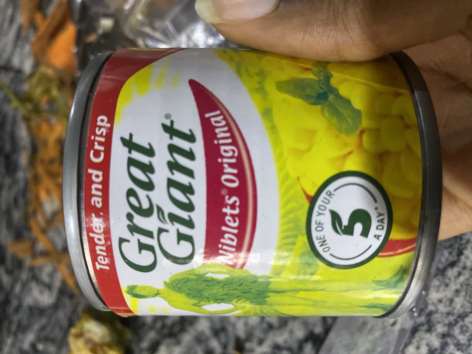 The image shows a close-up of a can of "Great Giant Niblets Original" corn. The can has a green figure on its label, and a red band with the words "Tender, and Crisp" printed in white. A circular green and white logo that says "ONE OF YOUR 5 A DAY" is visible on the lower part of the can. The background is blurred, but appears to be a countertop with some vegetables, possibly carrots, in the distance. A person's hand, with fingernails, is partially visible on the right side of the image. The scene is indoors, with a focus on the product packaging.