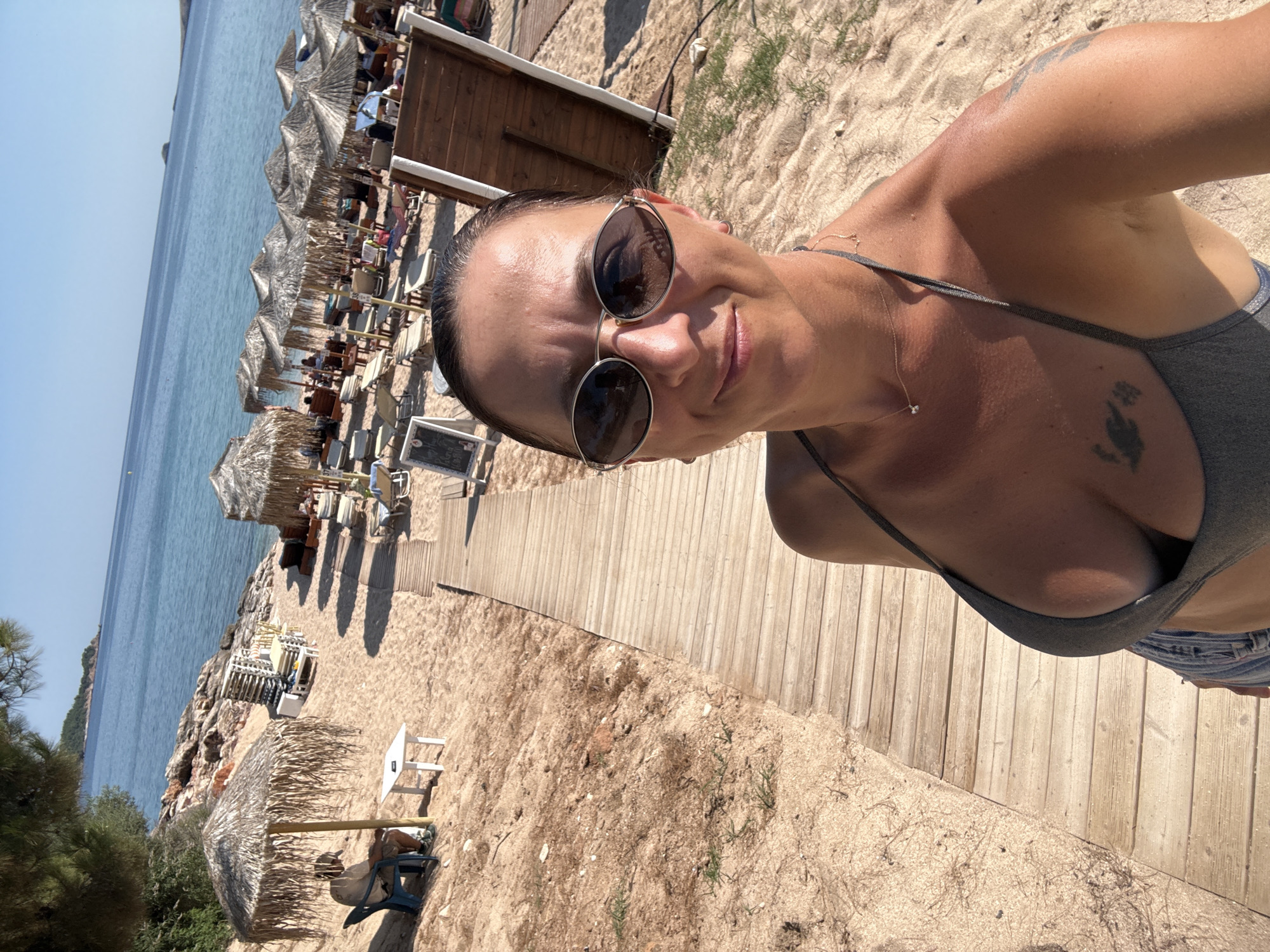 In a selfie taken on a sunny day at Potos Beach in Potos, Greece, a woman with dark hair pulled back and wearing sunglasses and a grey bikini top stands on a wooden boardwalk. The boardwalk stretches into the distance, flanked by sand, beach chairs, and straw umbrellas. The turquoise sea and a clear blue sky form the backdrop. In the background, other beachgoers are visible relaxing under umbrellas and on lounge chairs. The sand is light brown and appears fine-grained. The water is calm and inviting. A few trees are visible on the rocky shoreline to the left. The overall impression is one of a peaceful and idyllic beach scene.
