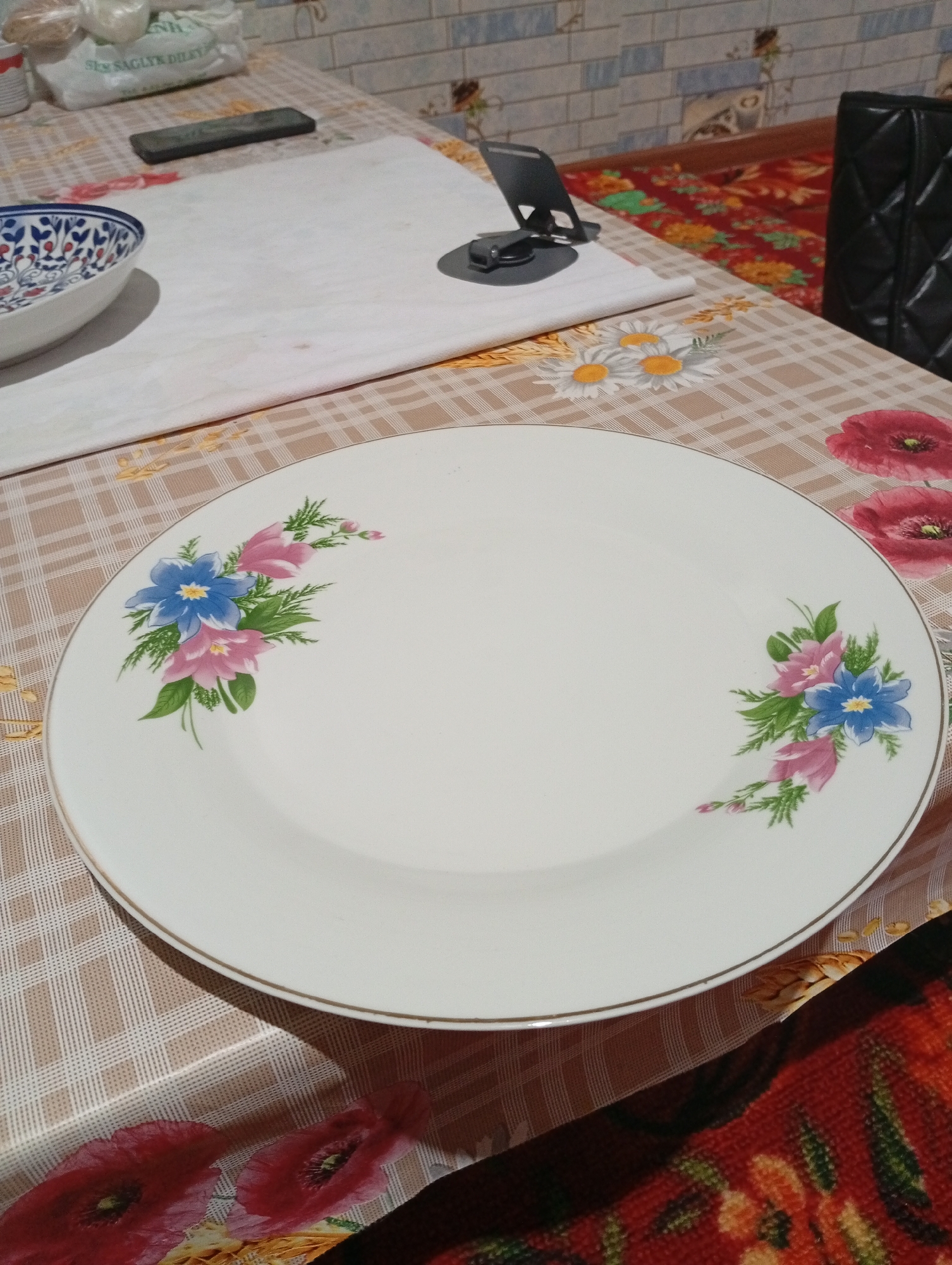 A white, round plate with a floral design is centered on a table covered with a checkered tablecloth. The floral design consists of pink and blue flowers with green leaves. To the left of the plate is a decorative bowl with a blue and white pattern. In the background, a portion of a tiled wall is visible. A black handbag and a mobile phone on a stand are also present on the table. The image appears to be taken indoors, possibly in a dining area. The specific location within Seýdi, Turkmenistan, cannot be determined from the visual information provided.