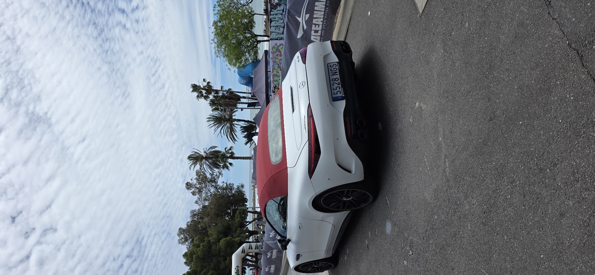 The image captures a white Mercedes-Benz AMG SL convertible with a red top parked on an asphalt road in Benicàssim, Spain. The car's license plate number is visible: 5528 NGS. Palm trees and other greenery line the background along with sky and clouds. Event banners are visible in the background for "OCEANMAN", suggesting an event is taking place in the area. The sky is overcast.