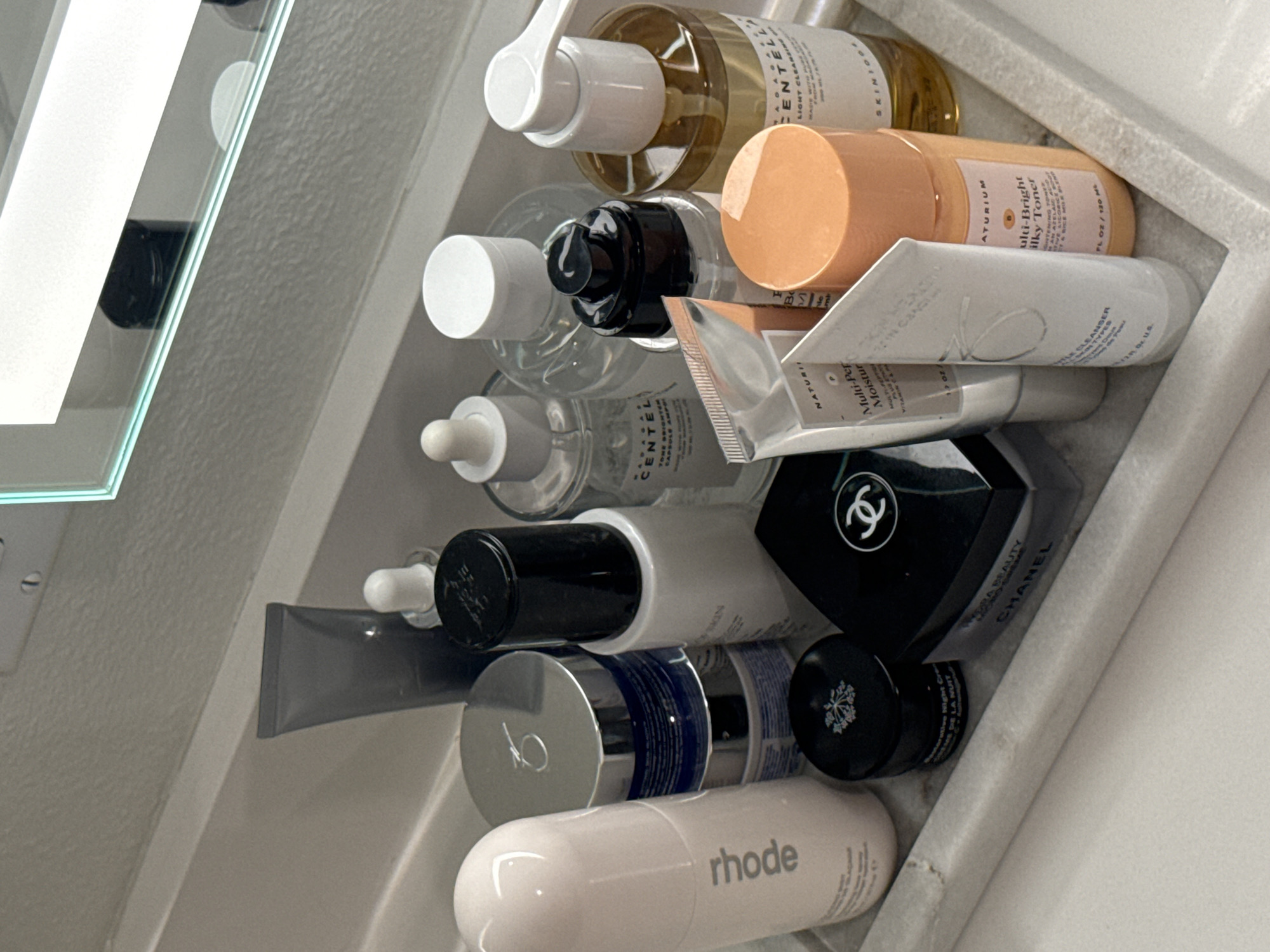 An overhead view captures a collection of skincare products neatly arranged within a white, textured organizer or shelf. The items, primarily bottles, tubes, and jars of various sizes, are positioned at an angle, suggesting a close-up perspective into a personal care space like a bathroom cabinet or vanity. No individuals are present, and no specific activity is occurring.
Notable brands and product types are visible:
- Clear bottles contain products like "SKINGOOD CENTELLA LIGHT CLEANSING" and "CENTELLA TONE BRIGHT CAPSULE AMPLE".
- An orange-peach bottle is labeled "NATURIUM Multi-Bright Silky Toner BRIGHTENING TONER".
- A white tube features "GENTLE CLEANSER".
- A silver-capped tube mentions "NATURIUM Multi-Peptide Moisturizer VITAMIN C".
- A black container displays "CHANEL HYDRA BEAUTY CHANEL".
- A small black jar has text indicating "Reparative Night Cream CREME REPARATRICE DE LA NUIT".
- A white, elongated oval bottle at the bottom clearly shows the brand "rhode".
The color palette is dominated by white, clear plastic, black, and soft peach/amber tones, with metallic accents. The items are tightly packed, indicating a well-utilized storage area. No specific time of day or weather conditions can be inferred from the image.