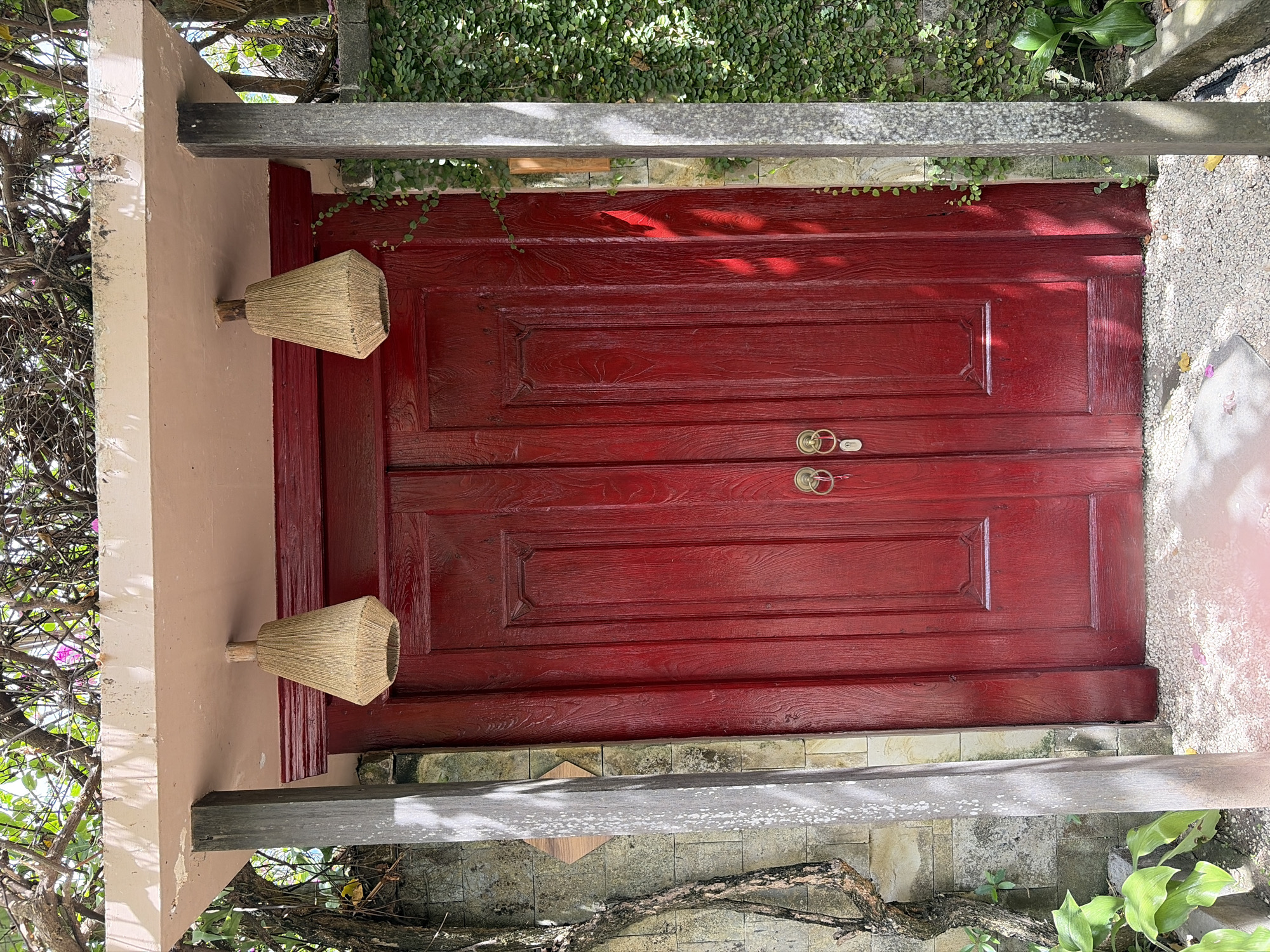 This image depicts the exterior of a building in Mengwi, Indonesia. The focal point is a pair of vibrant red double doors with ornate brass handles. The doors are flanked by two concrete pillars, and above them, a light-colored overhang shelters the entrance. Hanging from the overhang are two rustic, woven lampshades. The surrounding environment is lush and natural, with dense foliage and vines climbing the stone walls. The ground is covered in small pebbles, with some plants and branches visible at the edges of the frame. The scene suggests a tranquil and somewhat secluded entrance, possibly to a residence, restaurant, or boutique hotel.