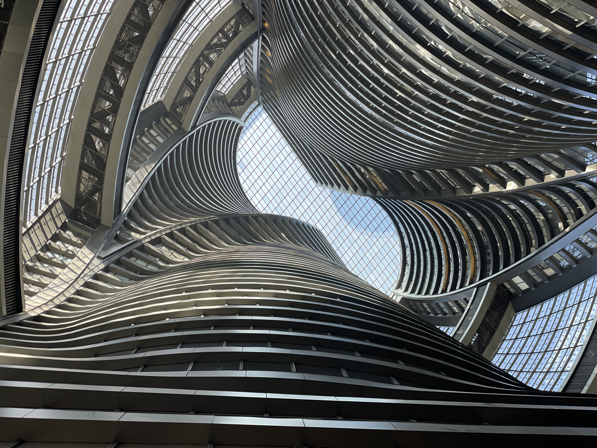 A low-angle shot within a modern architectural space, likely an atrium or lobby. The photograph is taken looking upwards. Curved, horizontally-layered structures rise and converge towards a grid-patterned glass ceiling that reveals the sky. The building materials appear to be metal and glass. The scene is well-lit, with light filtering through the ceiling. The location is Fengtai District, China. The building appears to be modern. No people are visible.