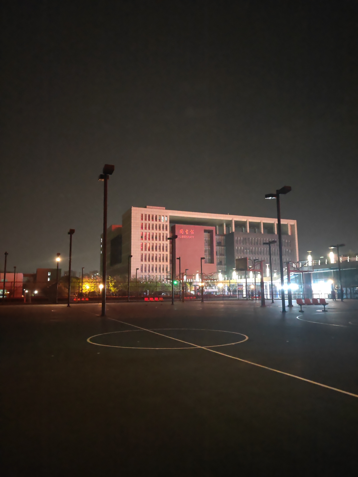 The image captures a night scene in the Jiangning District, China. In the foreground, there is a dark, paved sports court with white lines and a circle painted on it. Several tall light poles are interspersed around the court.
In the background, there is a large building with the word "LIBRARY" written in English, alongside Chinese characters. The building has a modern design, and the lights from inside illuminate some sections of it. The building is the Jiangning Campus Library of Hohai University. The sky above is dark and overcast.