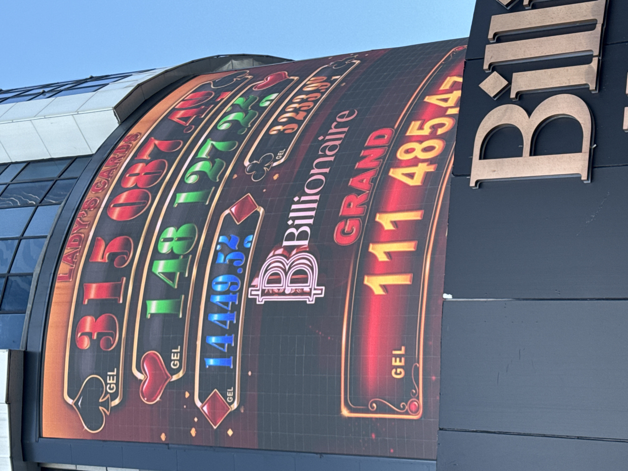 The image shows a large, curved sign attached to a dark gray building. The sign mimics the design of a slot machine, displaying various jackpot amounts in the Georgian Lari currency (GEL). The names of the jackpot are "Lady's Cards", "Billionaire Grand" and the names contain colored suits of cards. To the right of the sign is the beginning of the word "Billionaire" in large, gold-colored block letters. The building is located in Batumi, Georgia.