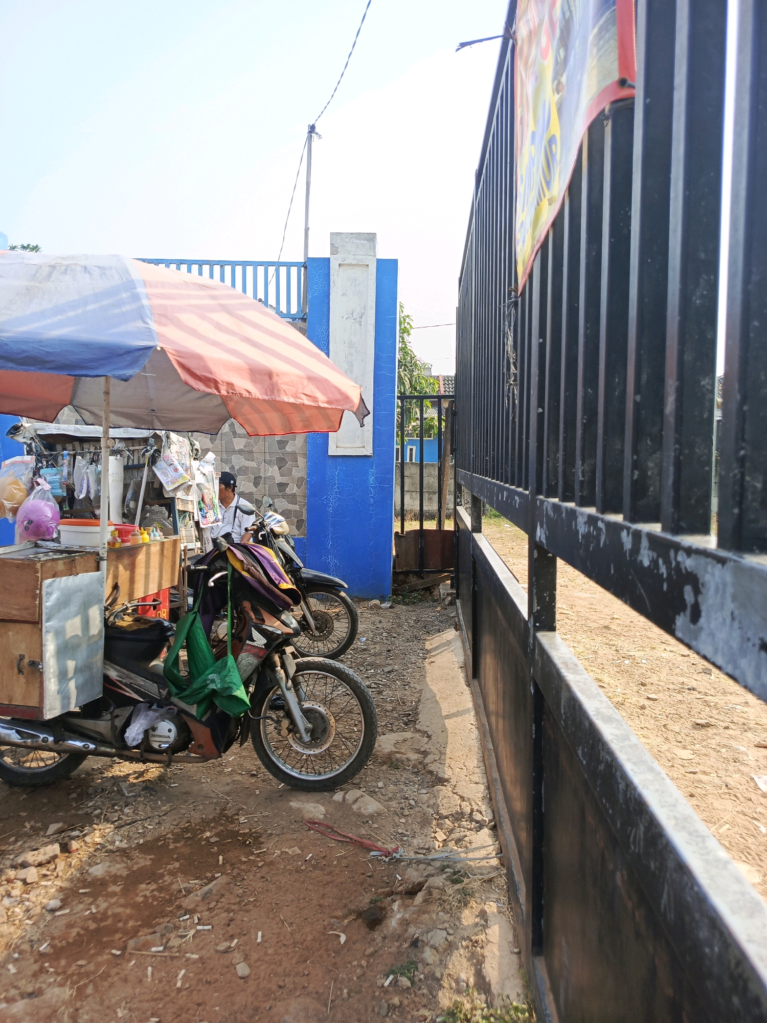 The image shows a street scene in Jati Mulya, Indonesia, with a focus on a small food cart. A man in a white shirt and black cap is seated on a motorcycle in front of the cart. The cart has a wooden structure with metal accents and is covered by a colorful umbrella. Various items are displayed on shelves behind the cart, including packaged goods and what appear to be food items. Two motorcycles are parked next to the cart. The background includes a blue building with a white pillar and a black metal fence. The ground is dirt and gravel, with some scattered debris. The overall impression is of a humble roadside stall operating in a possibly semi-urban or residential area.