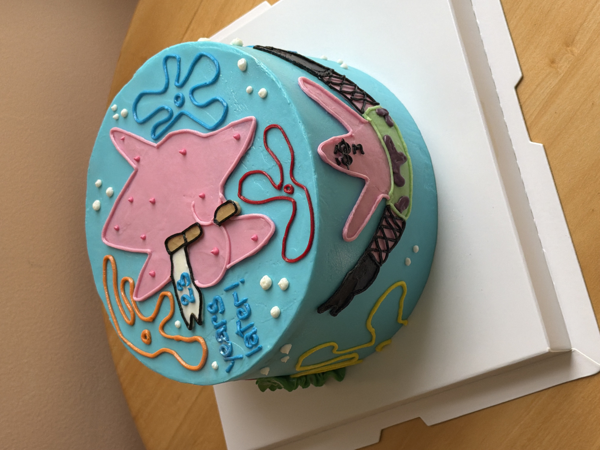 The image shows a round cake decorated with images of Patrick Star from Spongebob Squarepants. The cake is light blue with white pearl sprinkles. On the top, there's a pink Patrick Star figure holding two sticks with the writing, "23 years later!". A blue sea flower and wavy orange sea weeds, and one red. On the side, there's another Patrick Star figure wearing a green and purple outfit and black crossed arm bands. A yellow sea weed can be seen in the corner. The cake is on a square white cardboard base. The cake and the base are on a wooden table. The location of the cake cannot be determined from the image alone. Therefore, the city of Tel-Aviv, Israel, cannot be confirmed.
