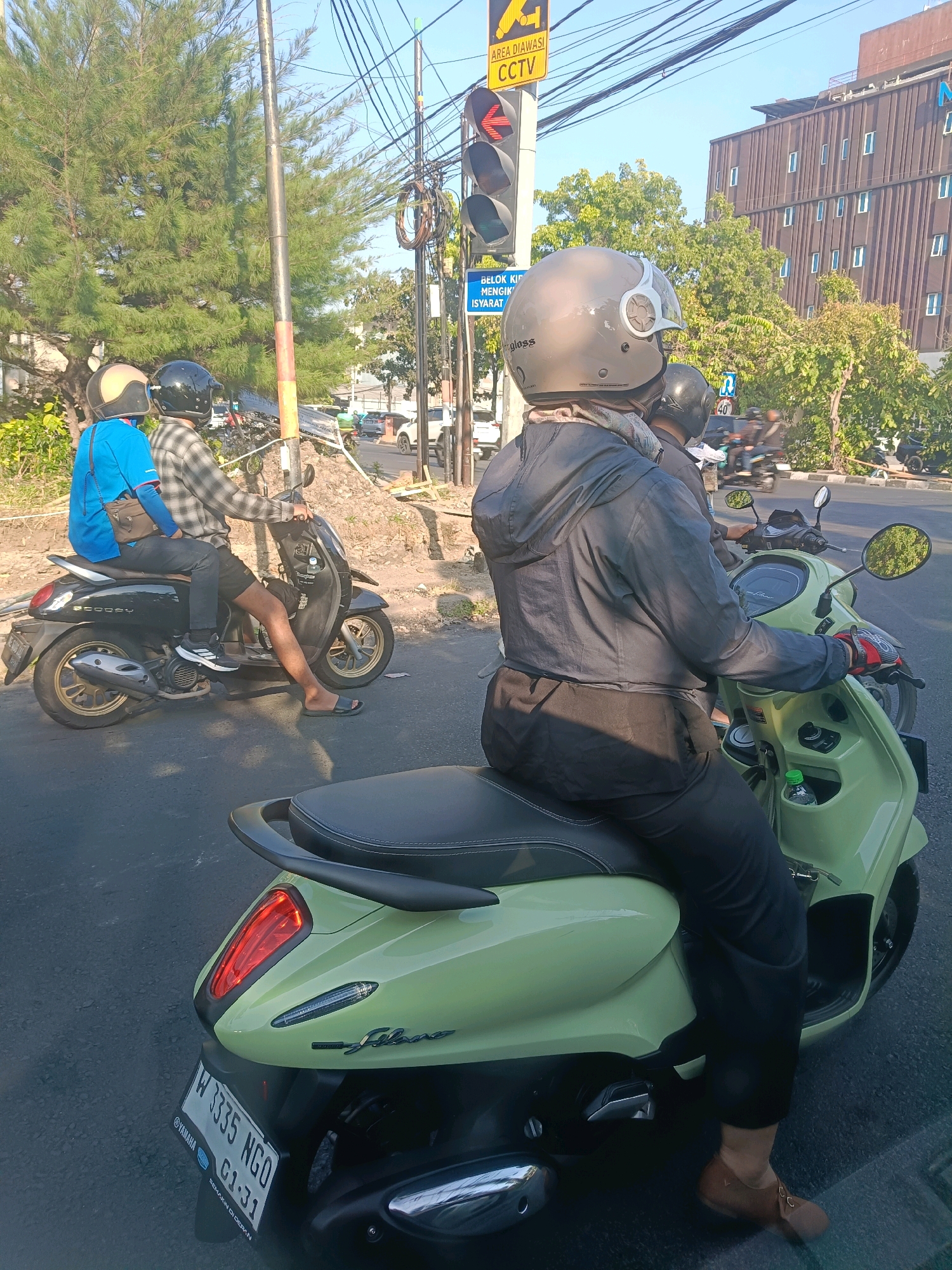 The image captures a street scene in Surabaya, Indonesia, at an intersection with traffic lights. In the foreground, a person wearing a grey jacket and black pants rides a light green Yamaha Filano scooter, their right hand with a red and black glove on the handlebar. The scooter has a license plate that reads "W 1335 NGO G1-31". Behind this scooter, another black scooter, a Honda Scoopy, is visible carrying two individuals. The person in front, wearing a blue shirt, has a brown shoulder bag. The person in the rear wears a plaid shirt and black shorts, with one leg exposed.
The environment is characterized by a clear blue sky and a street with asphalt paving. Trees line the street, and in the background, a modern building with a patterned facade stands. Electrical wires and traffic signals are prominent features of the urban setting. A yellow CCTV warning sign is mounted on a pole near the traffic lights. The overall impression is a typical day in a bustling Indonesian city.