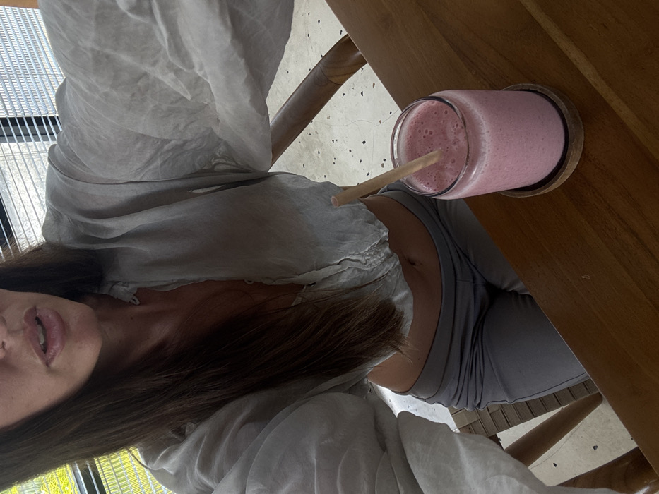 A first-person perspective shot captures a woman seated at a wooden table, enjoying a pink smoothie with a straw. Her attire includes a light-colored, slightly sheer blouse and a gray top. The composition is tilted, showcasing her lips and part of her face. Sunlight streams in from a nearby window.