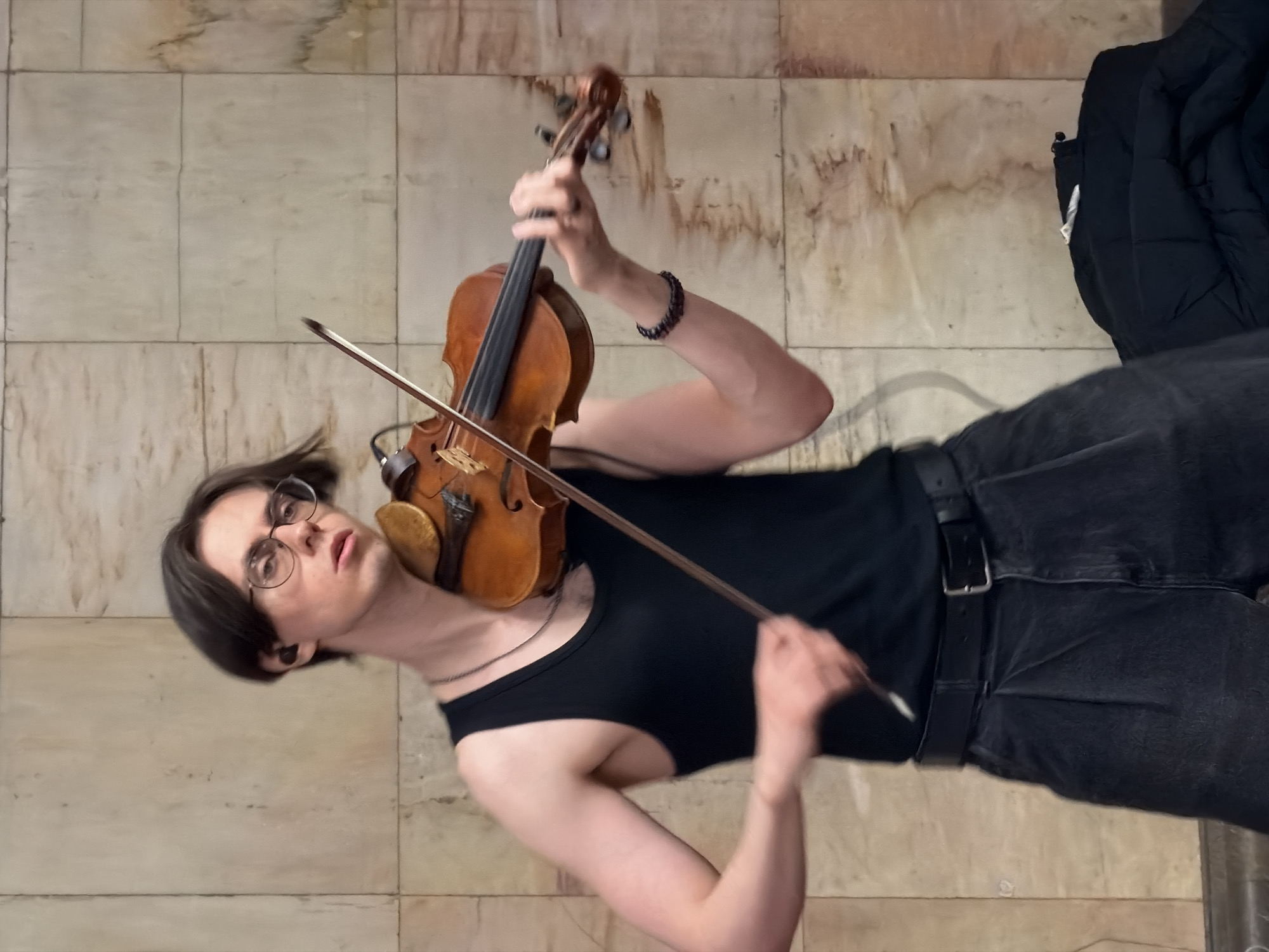 A young adult plays the violin while standing on a beige marble floor. The person is wearing round glasses, a black tank top, black jeans with a black belt, and a bracelet. A black jacket is on the floor nearby. The location appears to be a public space with a marble floor. There is no definitive indication in the image that the location is Moscow, Russia.