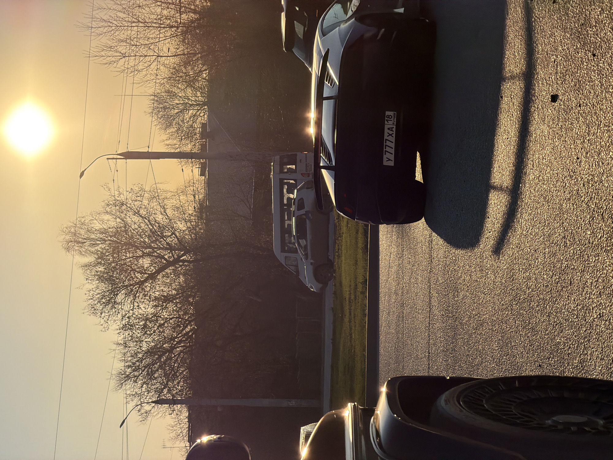 The image captures a street scene at sunset in Izhevsk, Russia, featuring multiple parked cars. The license plate of a dark sports car reads "Y 777 XA 18 RUS." A light-colored van is parked further down the street. Bare trees line the street, silhouetted against the bright sun. A streetlight is visible among the trees. The road surface reflects the warm glow of the setting sun.