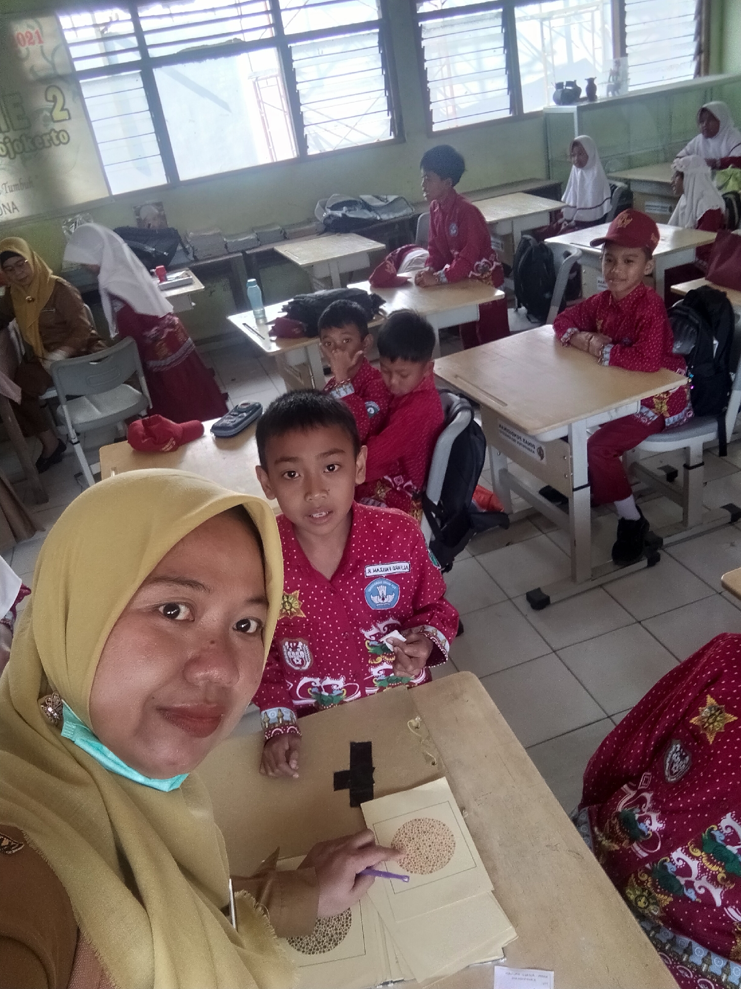 The image depicts an indoor scene, likely a classroom in Tretes, Indonesia, given the context provided. The room appears to be bright, with natural light streaming in from a large window with horizontal blinds. Several children, dressed in red and white school uniforms, are seated at wooden desks. One child in the foreground, wearing a red cap, looks directly at the camera. In the lower left corner, a woman wearing a yellow hijab and a blue face mask is visible, also looking towards the camera, taking a selfie. She is holding a piece of paper with small dots on it, and a purple pen. The desks are arranged in rows, and backpacks are visible on the floor and on chairs. The walls are painted a pale green. There are some posters and decorations on the walls. The floor is tiled. The overall atmosphere appears to be one of a school environment, possibly during a lesson or activity.