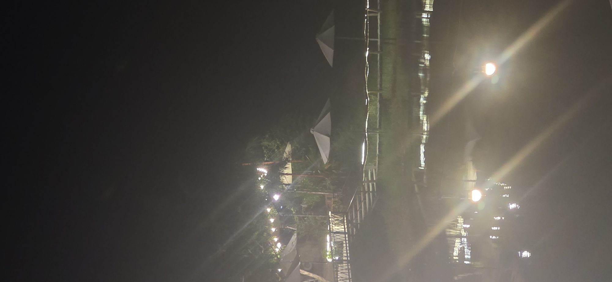 The image depicts an outdoor seating area at night, likely a restaurant or cafe, situated next to a body of water. The scene is illuminated by string lights and a few visible lamps, creating reflections on the water's surface. Several umbrellas are set up, suggesting areas for patrons to sit. A wooden walkway or staircase is visible, leading towards the seating area. The background features foliage and trees, indicating a natural or park-like setting. The time is night, and the overall atmosphere is calm and illuminated. There are no visible people or distinct actions taking place. The location is identified as being in Nha Trang, Vietnam.