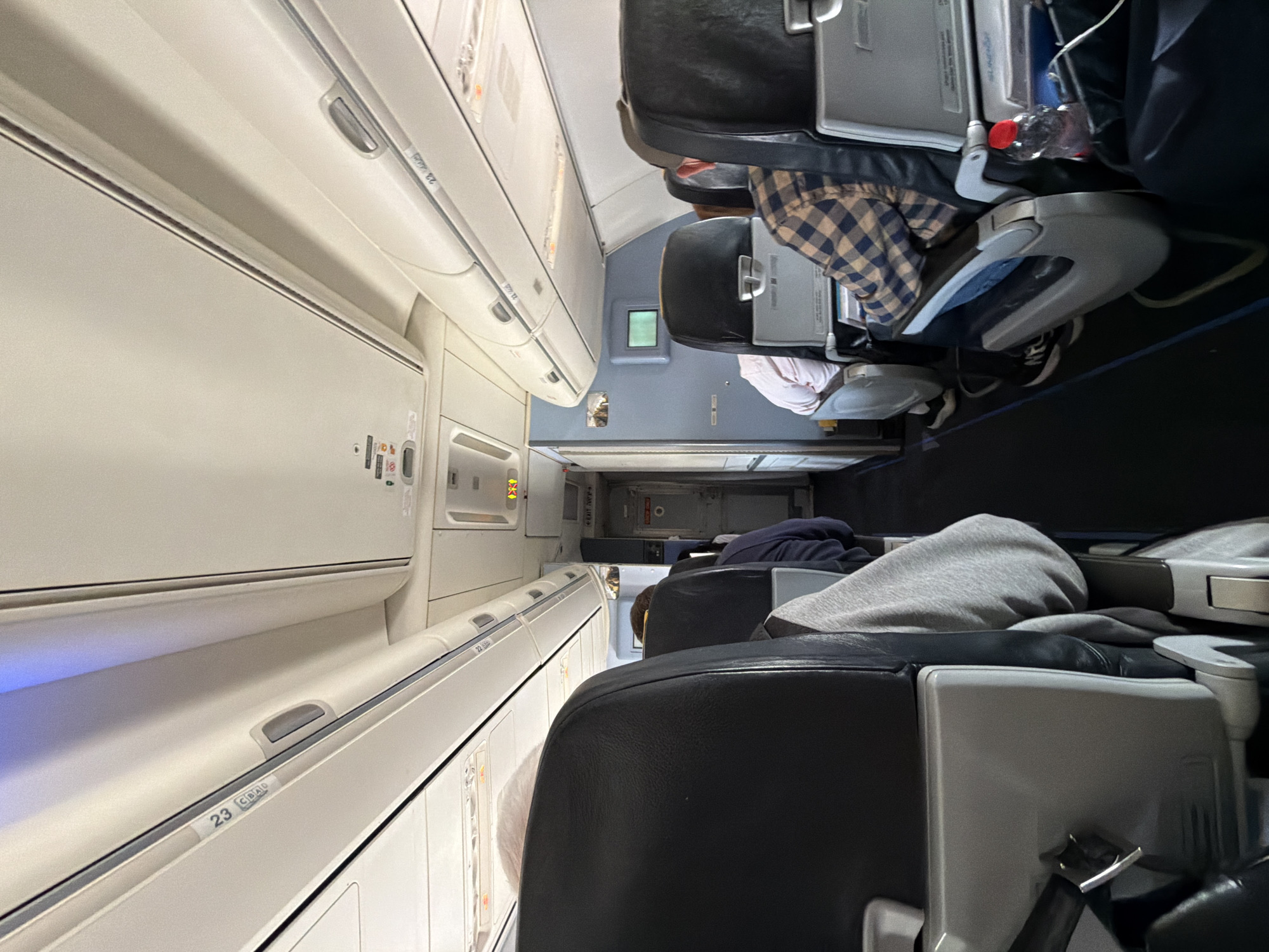 This media file captures the interior of an airplane cabin, rotated 90 degrees clockwise. The image looks down an aisle, with rows of passenger seats on both sides.
Several passengers are visible. In the middle-right foreground, a person is seated, wearing a brown and beige plaid shirt. A clear plastic water bottle is visible next to them. Further back, another passenger wears a light-colored (possibly pink) shirt. In the bottom-right foreground, the back of a black airplane seat is prominent, with a grey hooded sweatshirt draped over it, and another passenger partially visible. The seats are black with grey plastic components.
The overhead bins, typical of an aircraft, run along both sides of the aisle. They are light-colored, with handles and small information panels. Artificial cabin lighting illuminates the scene. The activity depicts passengers seated, likely during a flight or boarding/deplaning process.
Visible text on the overhead bins includes "23 CBAD" and "22 CBAO", indicating row numbers and seat letters. An emergency exit sign with the word "EXIT" and an arrow is partially visible further down the aisle. Small warning or instructional labels are also present on the cabin panels.
The overall setting is an aircraft cabin, consistent with travel, with the provided location context of Yagel, Israel, suggesting either the origin or destination of this journey. The time of day and weather are not discernible from within the cabin.