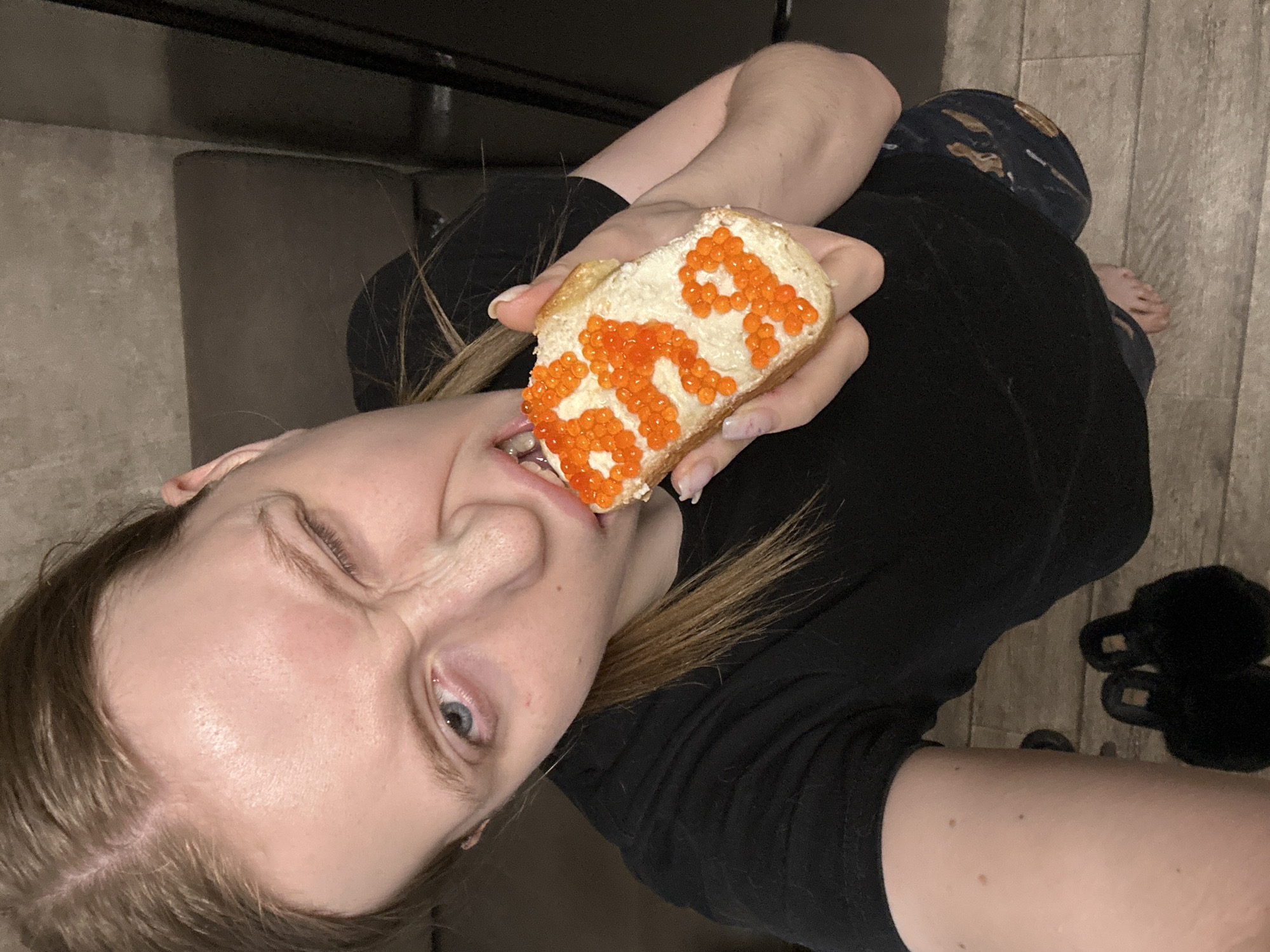 A Caucasian woman with blonde hair is holding a piece of bread covered with butter and caviar that spells "STR" toward the camera. She is winking and starting to eat the bread. She is wearing a black shirt and dark blue pants with a pattern. She appears to be indoors, based on the presence of what appears to be a couch, wood-look flooring, and black slippers on the floor. There is no evidence to confirm this image was taken in the city of Chaikovsky, Russia.