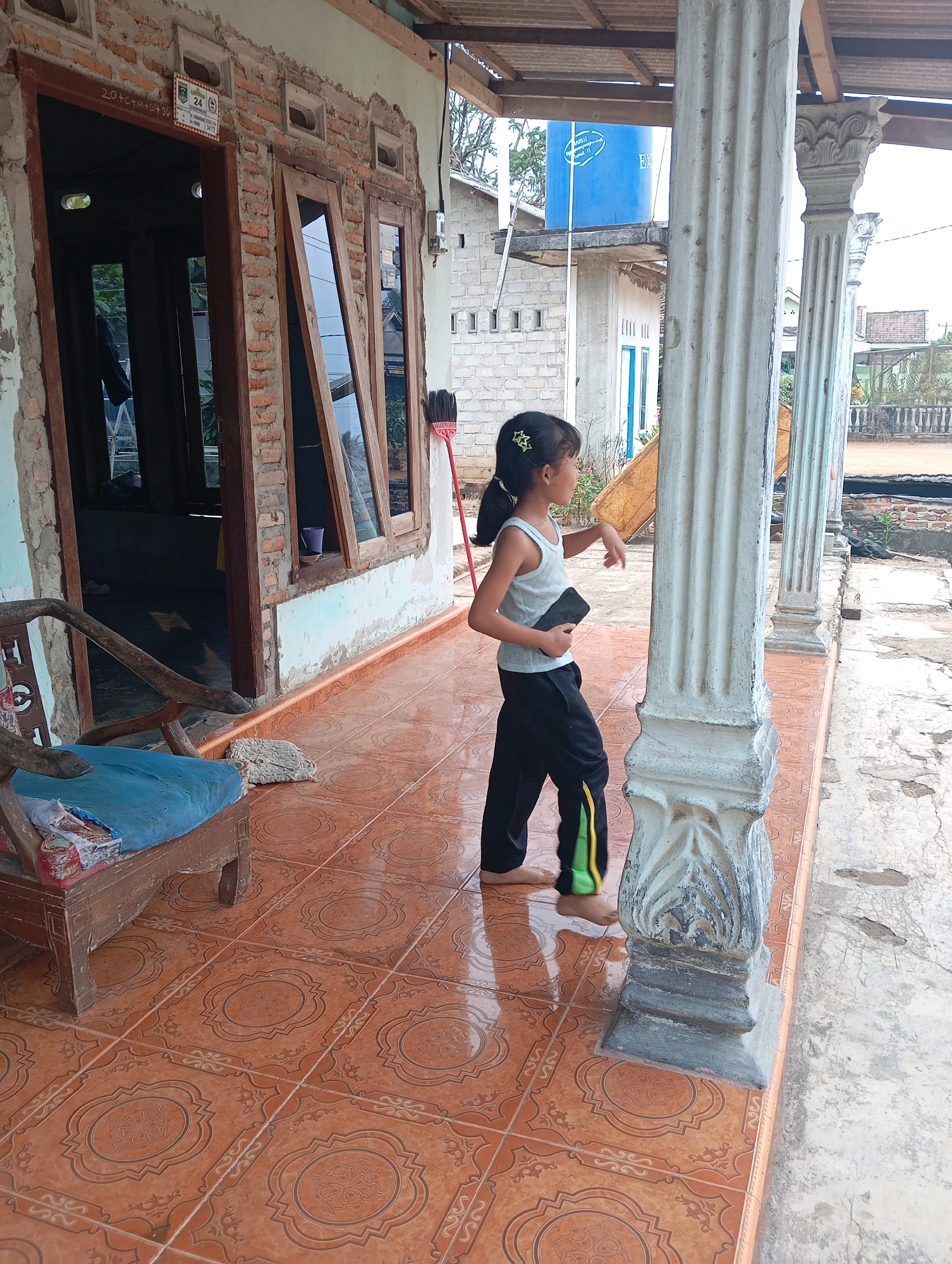 A young girl stands on a covered porch in front of a house. She is wearing a light-colored tank top and black pants with green and yellow stripes. She is holding a dark-colored rectangular object in her right hand. Her left hand is resting on a white pillar. The porch has orange tiled flooring with a decorative pattern. To the left of the girl, there is a wooden chair with a blue cushion. The house appears to be under construction or in disrepair, with exposed brickwork and peeling paint. A red broom leans against the wall to the left of the entrance. In the background, there are buildings, a blue water tank, and vegetation. The image is taken from a slightly low angle, looking towards the right. The overall scene depicts a residential setting in Leman Benawi, Indonesia.