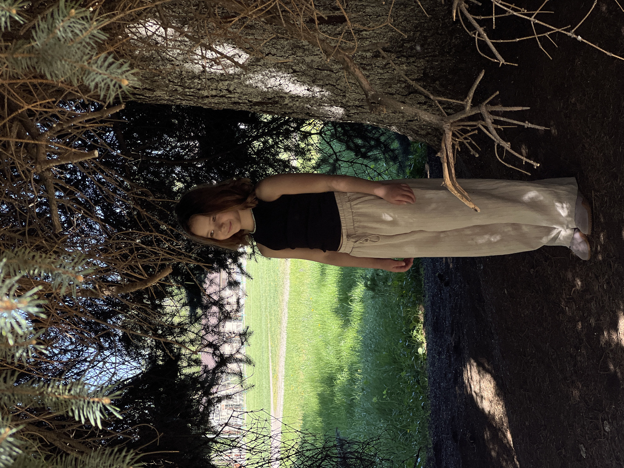 A girl stands beneath the canopy of a tree. The girl is wearing a black tank top and light tan pants. Part of a grassy lawn is visible in the background. The ground is dark and appears to be covered in dirt and tree debris. The location is likely Слобода, Russia.