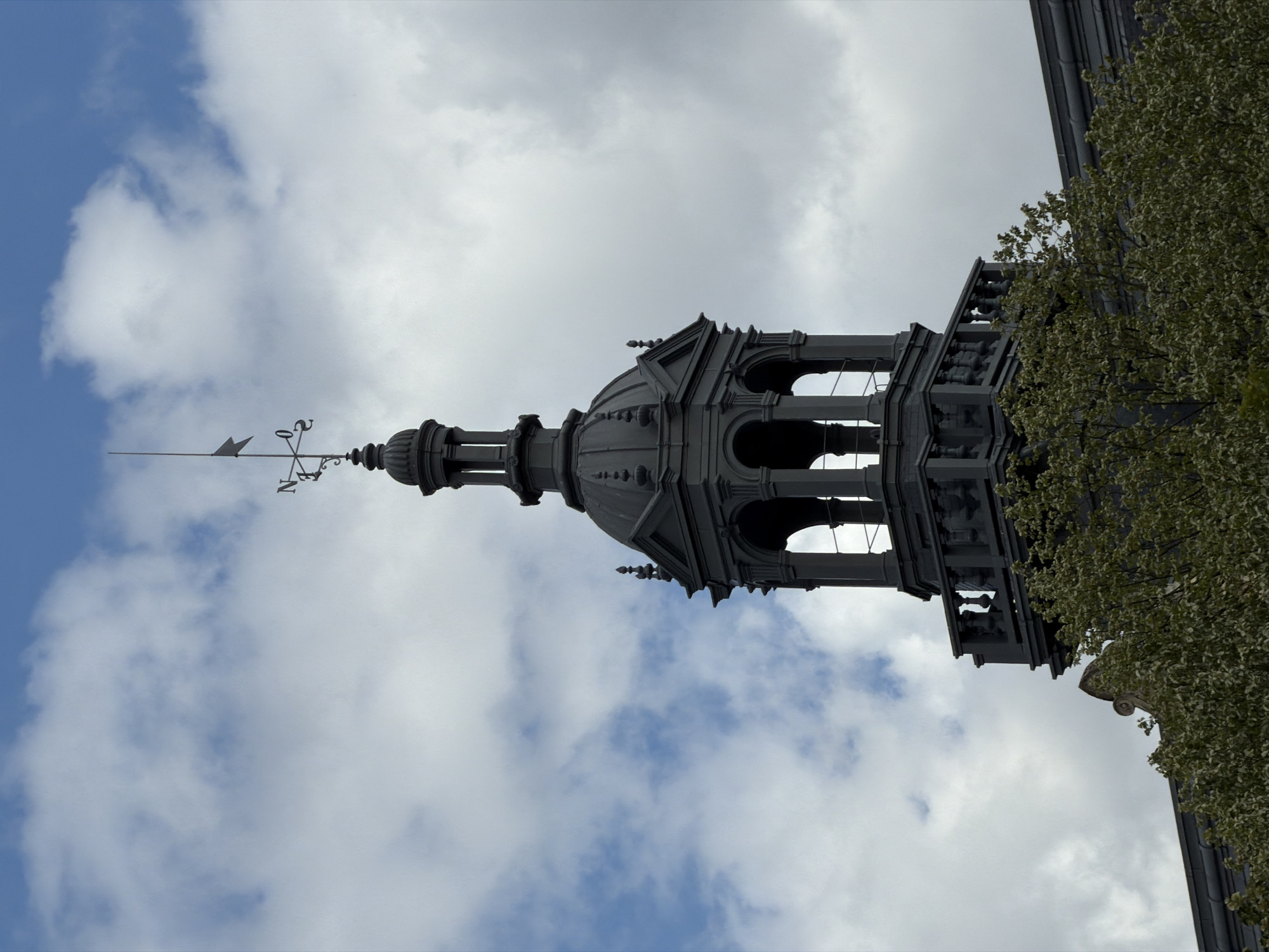 An image shows a view of a dark-colored, ornate clock tower with a weathervane on top, set against a cloudy blue sky. The tower has a domed roof and several tiers of columns and arches. A balustrade runs along a lower section of the tower. Green foliage from trees partially obscures the base of the tower and the roof of the building it is attached to. The weathervane is positioned on the very top of the tower, with the letters "N" and "S" visible, indicating directions. The overall impression is that of an old, possibly historic building. The image does not depict any people or specific actions. The location is specified as Ivry-sur-Seine, France.