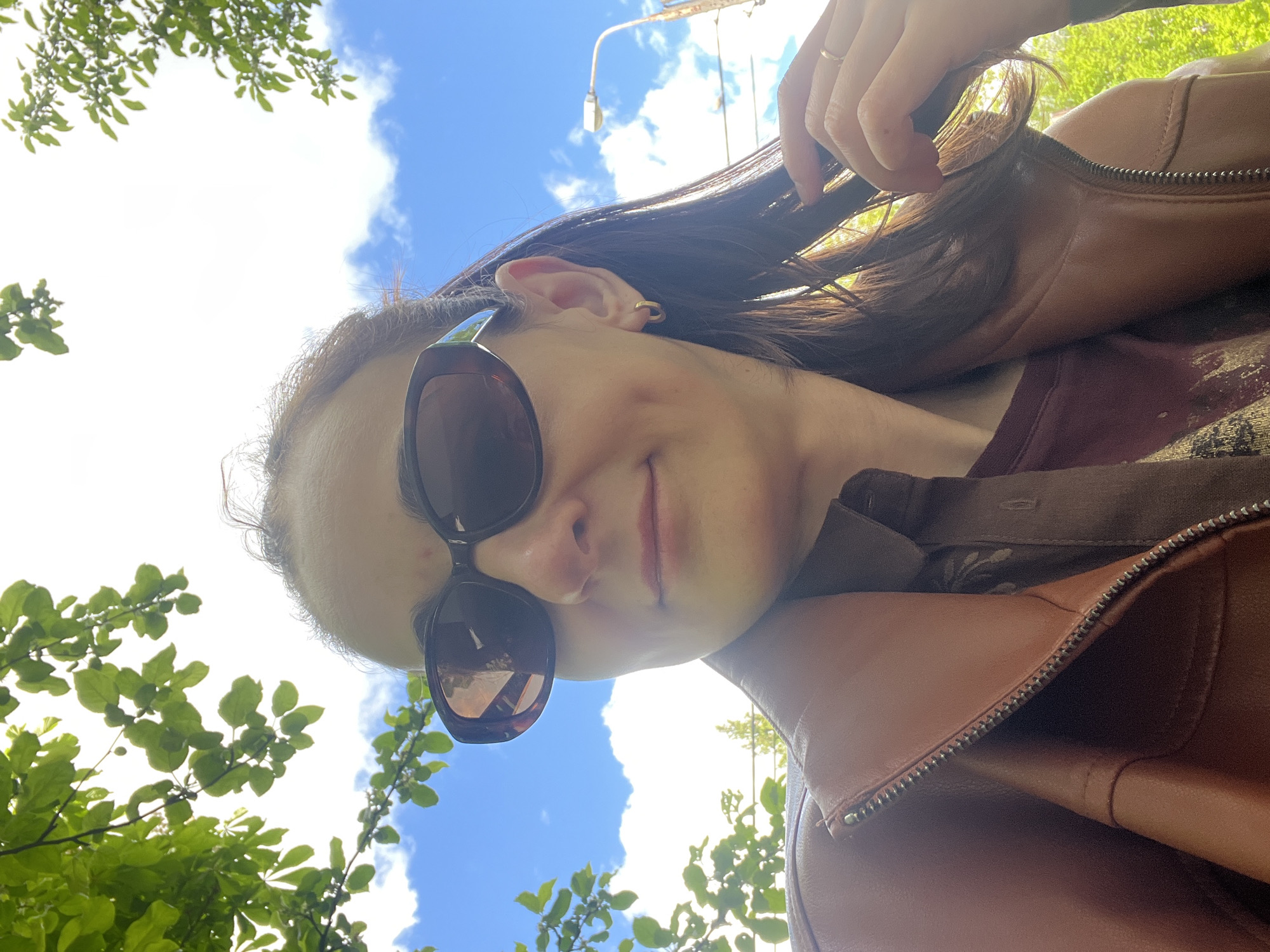 The image shows a woman in a brown leather jacket and sunglasses, with trees and a blue sky visible in the background. She is looking down at the camera and touching her hair with her right hand. A street light can be seen in the background. The location appears to be outdoors, possibly in a park.
