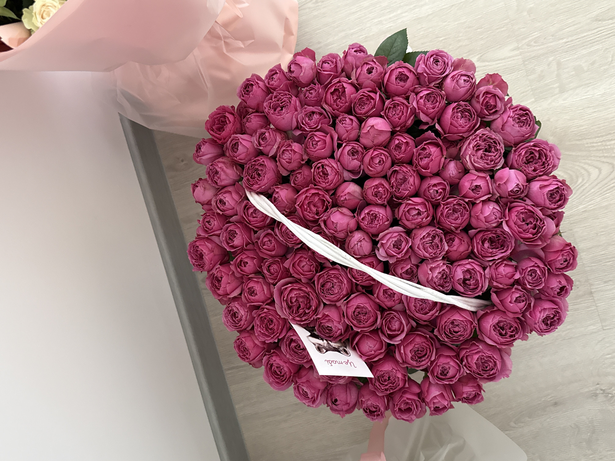 An overhead shot shows a large bouquet of deep pink, tightly packed roses arranged in a white basket. A small white card with a black and white drawing of a dog and some handwritten text in a cursive font is placed amongst the roses. The basket sits on a light-colored wooden floor next to a grey baseboard. In the background, soft pink tissue paper wraps a second, out-of-focus bouquet. The scene is illuminated by natural light, casting soft shadows. The location is described as Unknown, Unknown.