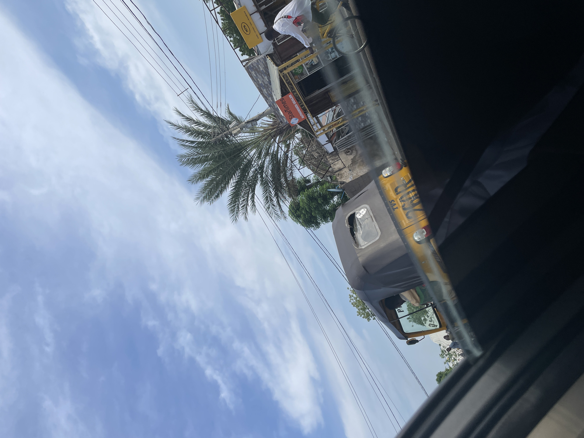 In Jalingo, Nigeria, an auto-rickshaw travels on a road under a partially cloudy sky. The auto-rickshaw is yellow with a black canopy and features the "TVS" logo on its side. A person is visible inside the auto-rickshaw. In the background, there is a palm tree, some buildings, and a person riding a bicycle. One building has a "MTN" sign and another has a "StarTimes AUTHORISED DEALER" sign. Electrical wires are strung overhead. The image is taken from inside a vehicle, with a dark interior visible in the foreground.