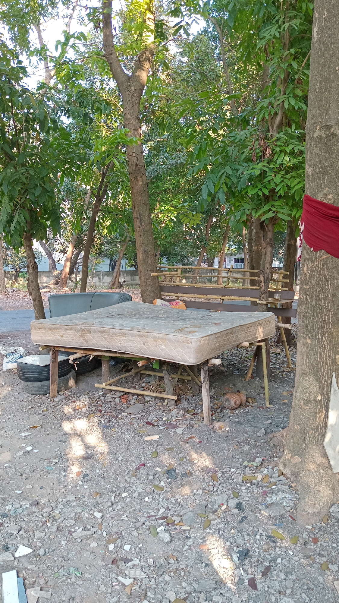 A mattress is placed on a makeshift wooden table outdoors. The mattress is situated on a dirt and gravel ground, surrounded by trees and vegetation. To the left of the mattress, there is a black car tire stack. In the background, a wooden bench and a couch are visible. The scene appears to be in a rural or semi-urban outdoor area, possibly in Randuagung, Indonesia, judging by the foliage and the style of the bench. The lighting suggests it is daytime.