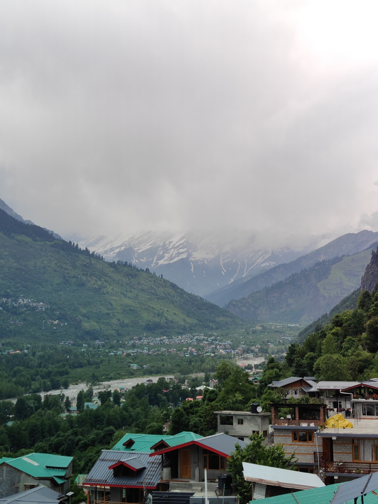 This is a landscape photograph taken from an elevated viewpoint overlooking the village of Vashisht, India. The image captures a valley nestled between lush green mountains, with snow-capped peaks visible in the distance under a cloudy sky.
In the foreground, a cluster of houses with distinctive green and grey tiled roofs are visible. These houses are built in a traditional style, some with multiple stories and balconies. The architecture suggests a mountainous region.
Further down the slope, the village of Vashisht itself is spread out, a dense collection of buildings with a variety of roof colors and styles. A river, appearing somewhat wide and turbulent, flows through the valley floor, lined by dense green trees. The mountains on either side of the valley are covered in vibrant green foliage, with some areas appearing to be terraced or farmed.
The upper portion of the image is dominated by the sky, which is heavily overcast with clouds. The light is diffused, creating a soft, atmospheric quality. The snow on the distant mountains is partially obscured by clouds, giving the scene a somewhat misty or hazy appearance.
The overall impression is one of a serene mountain village, possibly during or just after a period of rain, given the cloudy sky and the lushness of the vegetation. There are no people or animals prominently featured in the image. The focus is on the natural landscape and the built environment within it.