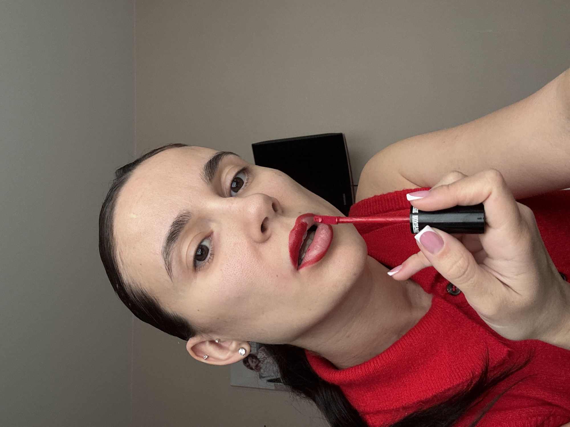 A woman is applying red lip gloss while wearing a red sweater. She has her hair pulled back and is wearing earrings. In the background is a grey wall and a black television. There is no indication in the image that it was taken in Chaikovsky, Russia.