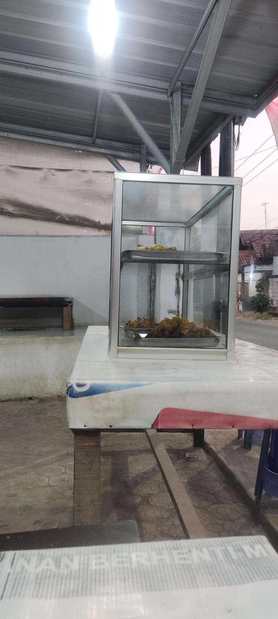 A glass and aluminum display case sits on a table outdoors, likely in the city of Getrakmoyan, Indonesia. The display case contains two trays of food, one with what appears to be whole chickens and the other with pieces of fried food, possibly chicken or fish, on skewers. The case is lit from above by a bright fluorescent light. The table is covered with a white tablecloth that has blue and red accents. In the background, there is a metal roof structure, a wall, and some buildings with tiled roofs, suggesting an outdoor food stall or eatery. The ground is paved with stone tiles. The overall impression is of a simple, functional setup for selling food in an open-air setting.