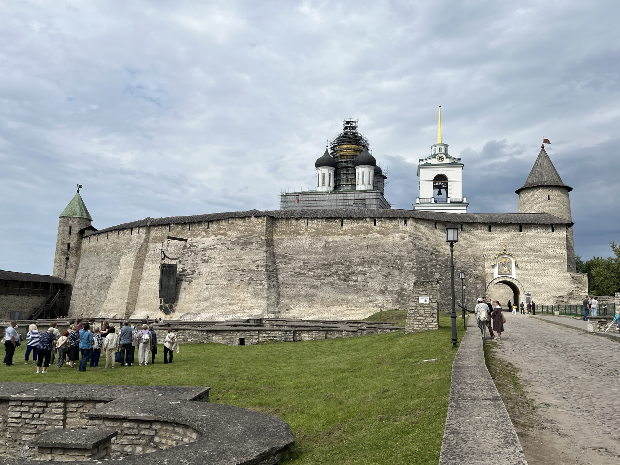 The image depicts the Pskov Kremlin in Pskov, Russia. The massive stone walls of the Kremlin dominate the foreground and midground, with a large green lawn in front of them. Several people, appearing to be tourists on a guided tour, are gathered on the lawn and walking along a cobblestone path leading towards an arched gateway in the Kremlin wall.
Several towers and buildings are visible atop the walls. On the left, a conical green-roofed tower with a flag is prominent. In the center, a large cathedral with multiple black domes, one of which is covered in scaffolding, is under renovation. To the right of the cathedral, a white bell tower with a golden spire stands tall. Further to the right, another stone tower with a conical wooden roof and a red flag is visible. The sky is overcast with a mix of light and dark clouds. The overall scene suggests a historical site being visited by tourists, with ongoing restoration work on one of the structures.