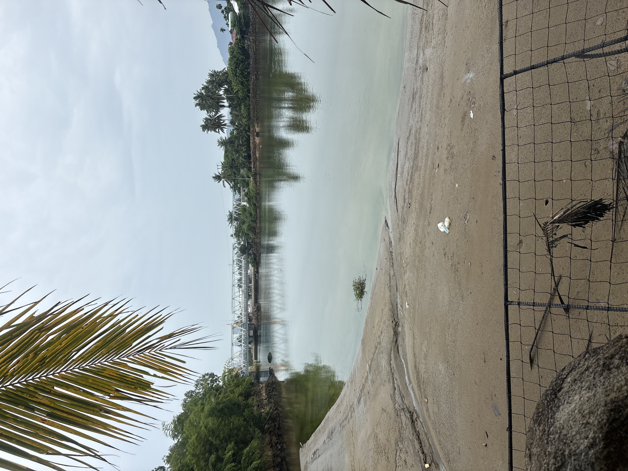 The image depicts a tranquil scene along a waterfront in Nha Trang, Vietnam. In the foreground, a sandy embankment slopes down to a calm, greenish body of water. A rough, concrete ramp is visible on the left, and a black metal fence runs across the bottom of the frame, partially obscuring a large, grey rock. Scattered pieces of white plastic debris are visible on the sand.
Across the water, a long, white truss bridge spans the waterway, with several vehicles and structures visible on it. Lush green trees line the opposite bank, interspersed with palm trees and some buildings. The sky is overcast with a pale grey hue. A frond of a palm tree hangs into the frame from the top left corner. The scene suggests a coastal or riverine setting within the city, potentially a less developed or industrial area given the presence of debris.