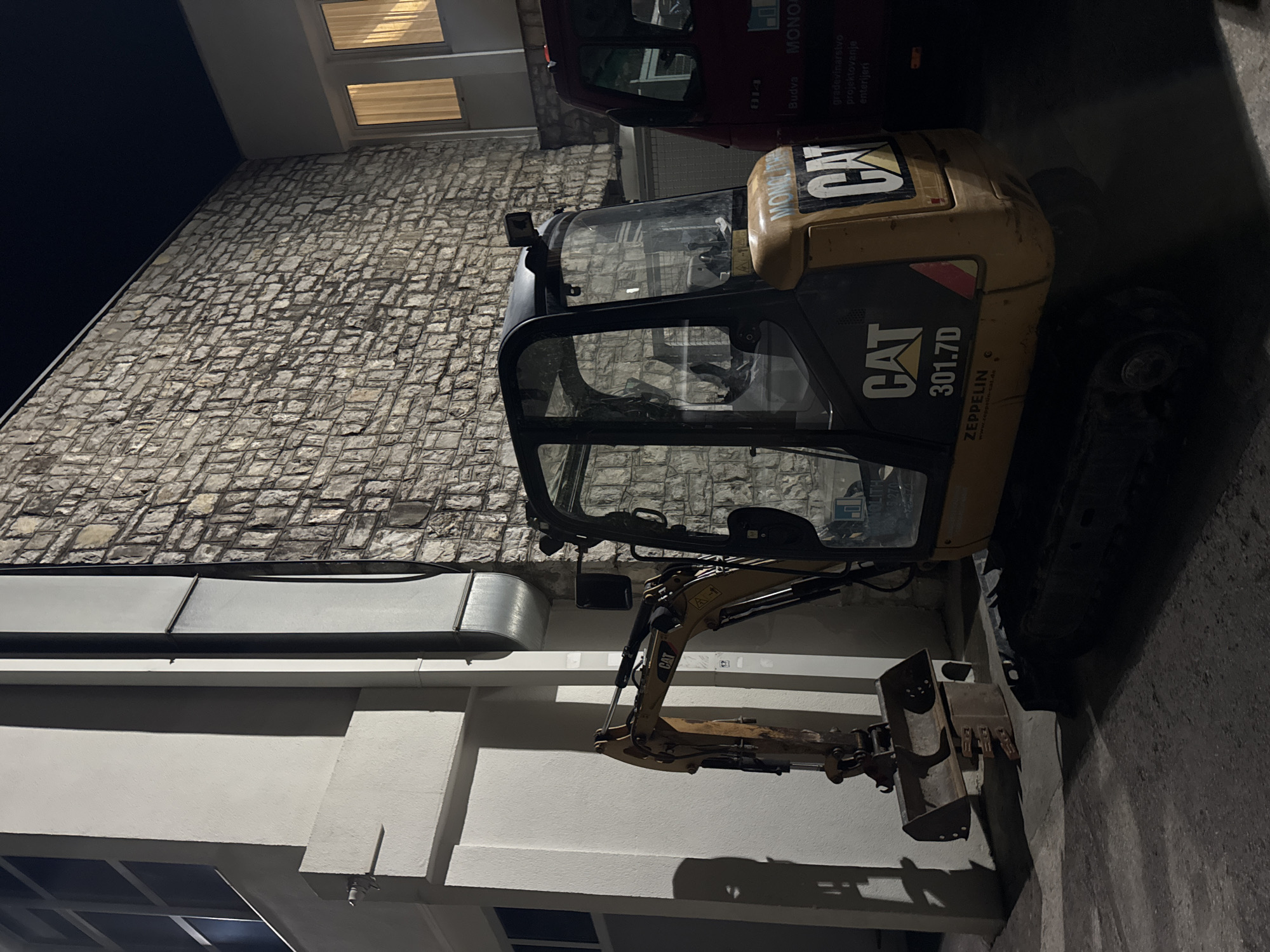 An excavator is parked at night in front of a stone building and a white building, possibly in the city of Boreti, Montenegro. The excavator is yellow and black, with the brand "CAT" visible on its side. Its arm is extended and its bucket is lowered to the ground. A red truck is parked to the right of the excavator. The environment appears to be a construction site or a storage area. The lighting is artificial, casting shadows on the ground and buildings. The sky is dark, indicating nighttime.