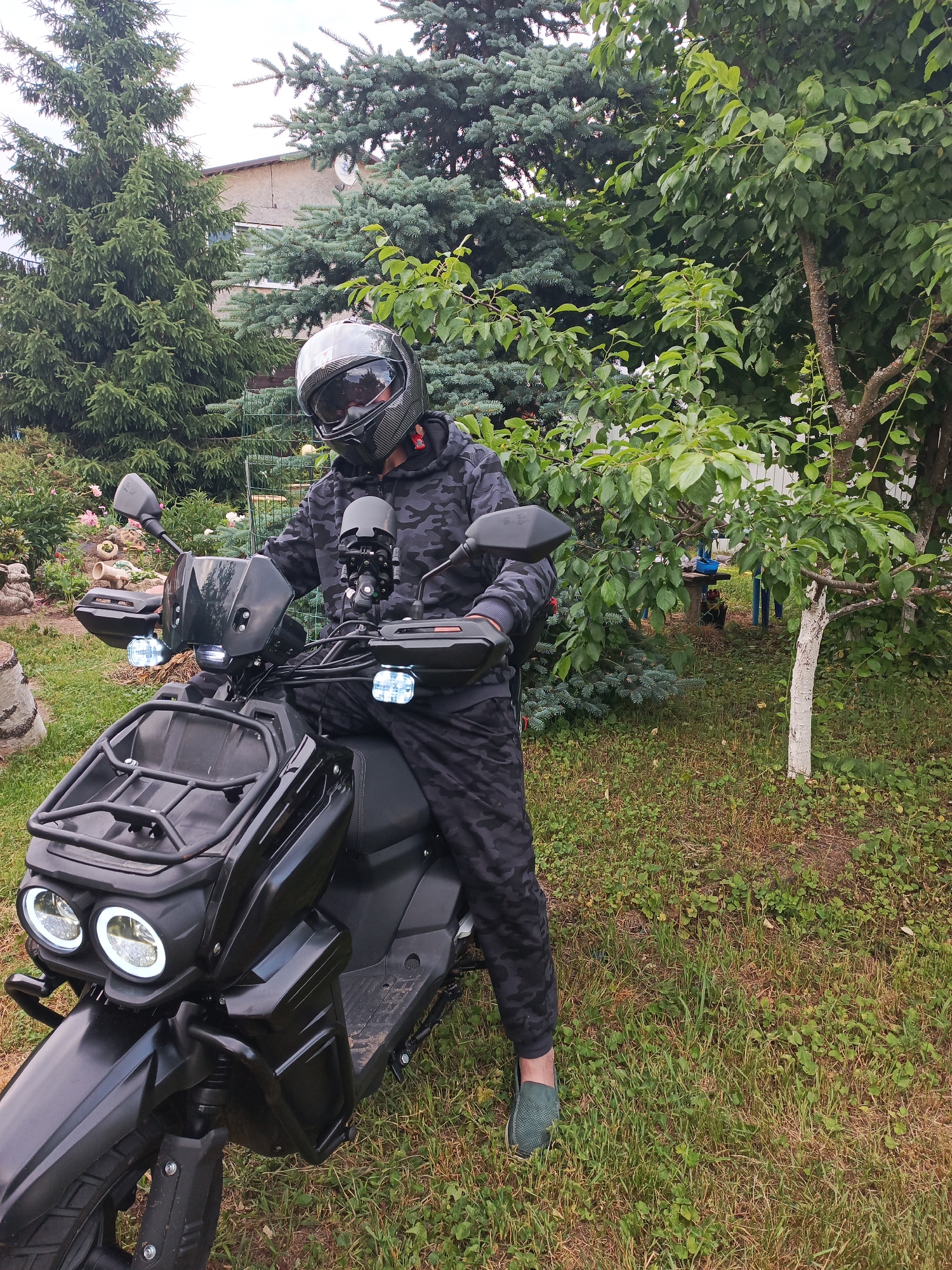 A person wearing a camouflage tracksuit and a black helmet with a reflective visor is sitting on a black scooter. The scooter has dual headlights and a front rack. The scene is outdoors, with a backyard environment featuring green grass, trees, and bushes. In the background, a two-story building and a fence are visible. The location is identified as Novoye, Russia.