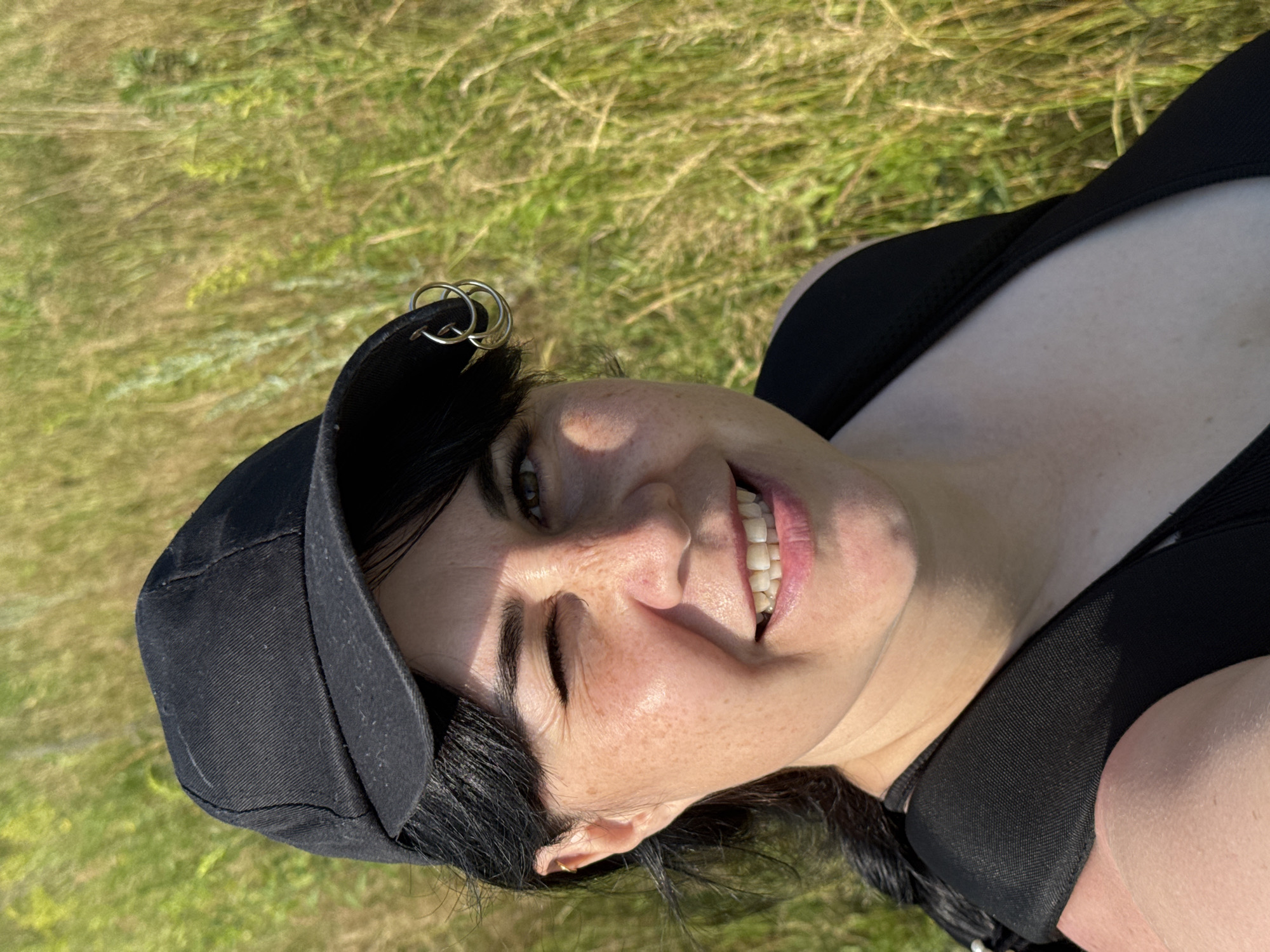 The image is a close-up, selfie-style shot of a woman outdoors. She is wearing a black baseball cap with silver rings attached to the side and a black tank top or sports bra. Her right eye is closed in a wink, and she is smiling, showing her teeth. She has dark hair and freckles on her face. The background is blurred and appears to be tall, dry grass, suggesting an open, natural environment. There are no identifiable landmarks or indications of the city of Izhevsk, Russia.