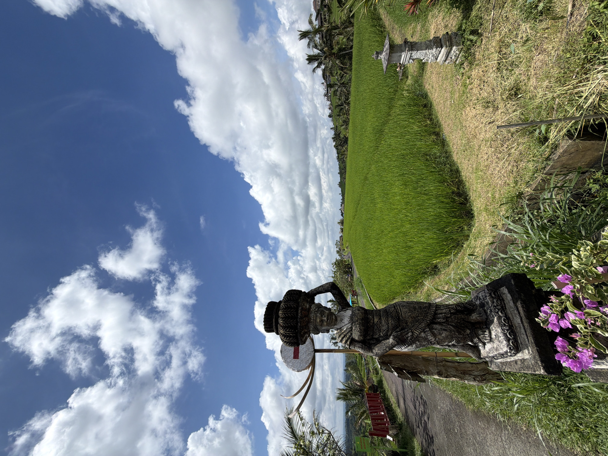 The image depicts a rice paddy landscape in an unknown location in Indonesia, taken on a clear day with a blue sky and scattered white clouds. A stone statue of a woman carrying what appears to be a stack of pots on her head is the focal point, positioned along a narrow paved path or road that borders the rice fields. The path is lined with vegetation, including some purple flowering plants. Further into the scene, lush green rice paddies stretch out, bordered by palm trees and other foliage. A second, smaller stone structure or shrine is visible further down the path alongside the rice field.