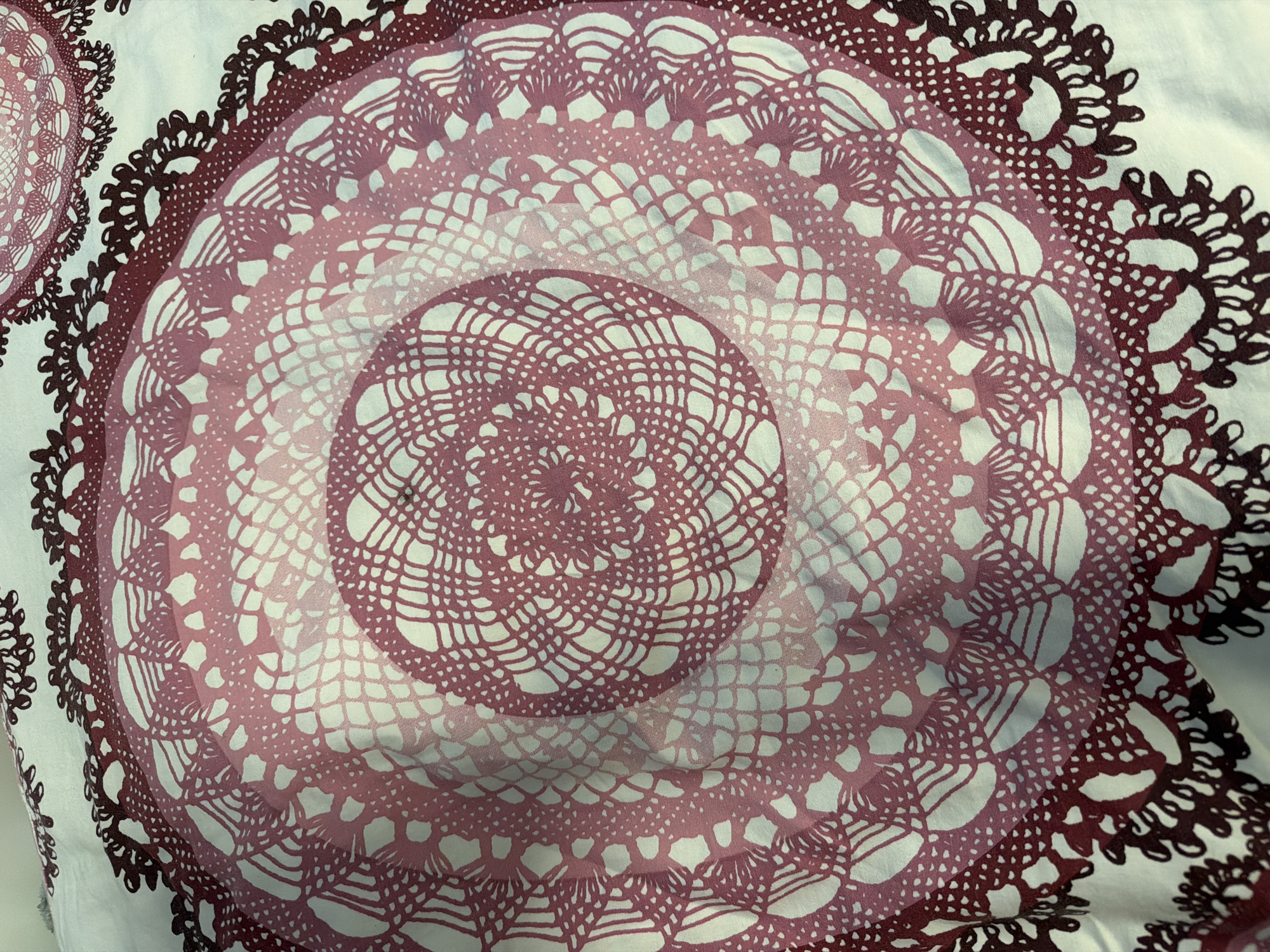 This is a close-up, overhead view of a fabric with a repeating circular, doily-like pattern. The pattern features concentric rings of intricate, lace-like designs. The colors transition from a deep reddish-brown at the outer edges to lighter shades of pink and white towards the center, creating a gradient effect. The design appears to be printed or dyed onto a white background. The fabric is slightly creased and appears to be a light material, possibly cotton or a blend. The lighting is neutral, suggesting an indoor setting without strong directional light. There are no people, animals, or specific setting details visible. The focus is entirely on the textile pattern itself.