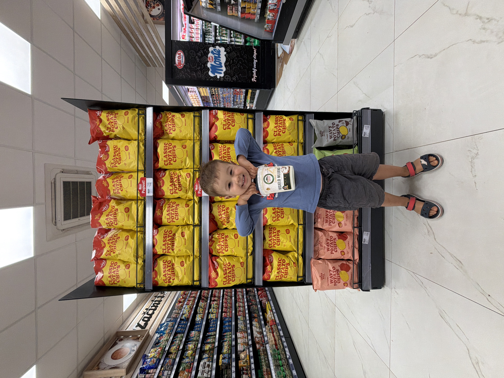 A young boy with a cheerful, wide smile stands in a brightly lit grocery store aisle, looking directly at the camera. He wears a blue long-sleeved shirt, dark shorts, and red and blue sandals, holding a white plastic container of "KISJELO MLIJEKO" (sour milk/yogurt) to his chin.
He is positioned in front of a tall display rack filled predominantly with yellow bags of "CLASSIC POTATO CHIPS SALTED," each weighing "70g." On the right side of the rack, there are also pink bags of "WAVY POTATO CHIPS PAPRIKA." In the background, to the left of the boy, shelves are stocked with various packaged items, including a section labeled "Zacini" (spices). To his right, a large black refrigeration unit for "MEGA PROMET" is visible, featuring the brand "Monte" and the slogan "Prijatelj vašeg džepa!" (Friend of your pocket!). The store features white tiled floors, white walls, and bright overhead lighting. A small air vent is visible on the wall. A sign for "Popust!" (Discount!) can also be seen. The boy's happy expression suggests a pleasant experience in this retail setting in Budva, Montenegro.