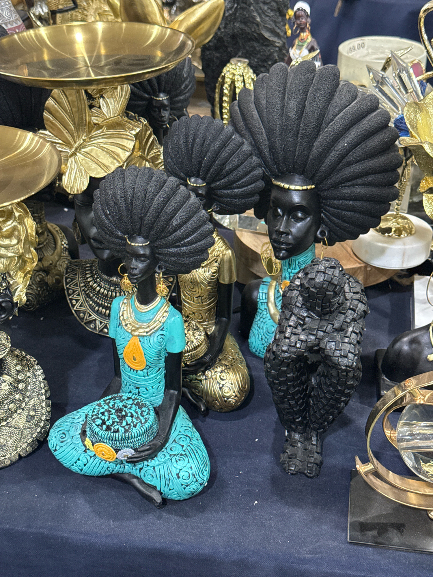 The image shows a collection of decorative items, likely for sale, displayed on a dark blue surface. In the foreground, a statue of a Black woman with a large, black afro hairstyle and wearing a turquoise dress with gold accents is kneeling. She is holding a round, decorative basket on her lap. To her right, another statue of a Black figure, this one with a more abstract, textured black surface, is hunched over. Behind these figures are more decorative items, including gold platters and intricate statues with large afros, some in gold and black. A small statue of a woman in traditional attire is visible in the background. The overall impression is of an eclectic display of African-inspired art and decor. The image does not contain any clues to suggest a location in Tbilisi, Georgia.