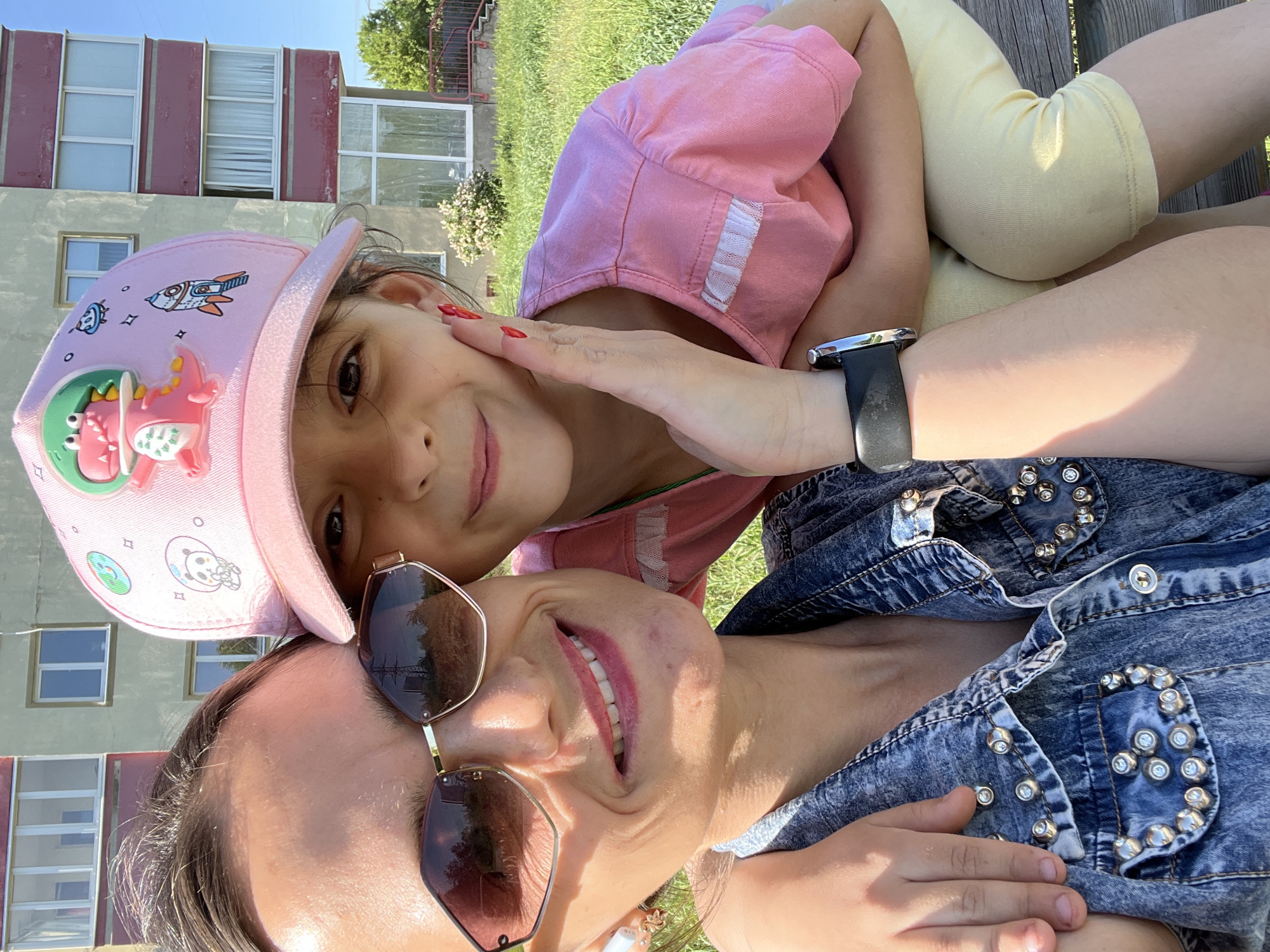 The image depicts a woman and a young girl, likely a mother and daughter, posing closely together in an outdoor setting. The woman is wearing sunglasses and a denim jacket adorned with metal studs, and the girl is wearing a pink hat and shirt with yellow pants. The girl's hand is gently touching the woman's face. The background includes a multi-story building with visible windows and green grass. The location is identified as the city of Chaikovsky, Russia.
