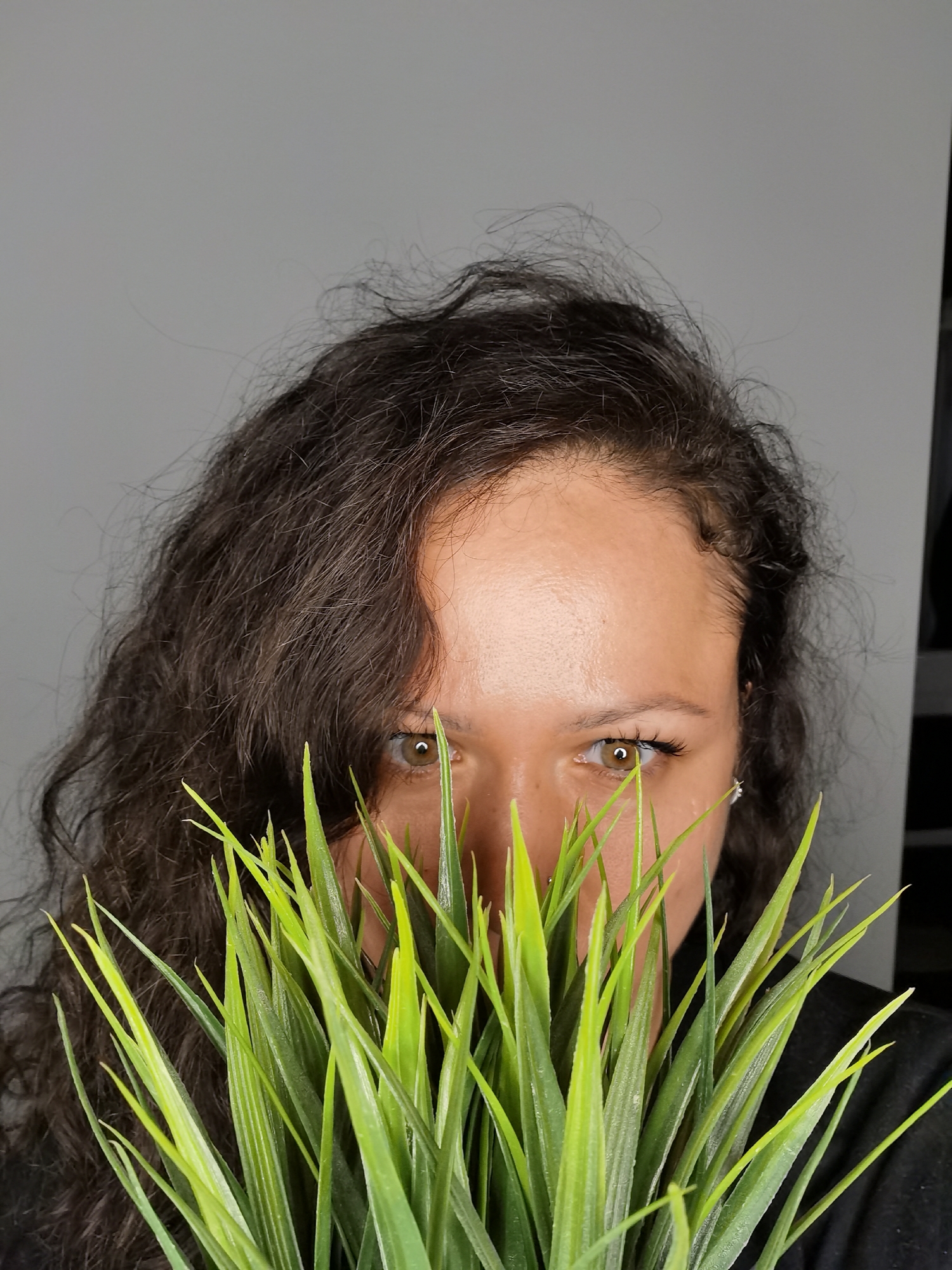 A person with curly dark hair and light brown eyes is holding a potted plant with spiky, green leaves in front of their face. The plant obscures the lower part of their face, leaving only the eyes and forehead visible. The person is wearing a dark-colored shirt. The background is a gray wall. This image provides insufficient information to determine if the scene is located in Chaikovsky, Russia.