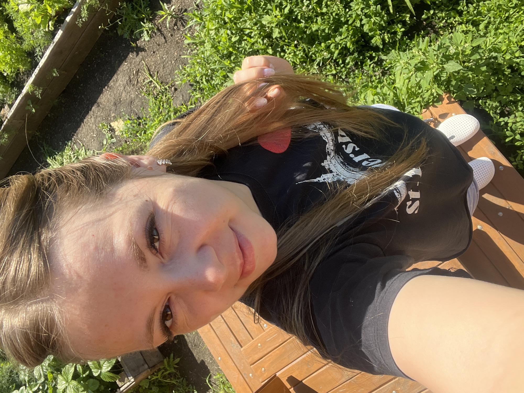 A young woman is taking a selfie outdoors, standing on wooden planks in what appears to be a garden. She has long brown hair styled in two braids that wrap around her head and a ponytail. She is wearing a black t-shirt with a graphic print and white sneakers. Her left hand is holding a strand of her hair. The background shows greenery and a raised garden bed. The lighting suggests it is a sunny day. The image does not provide any specific information to place it in the city of Shadrinsk, Russia.