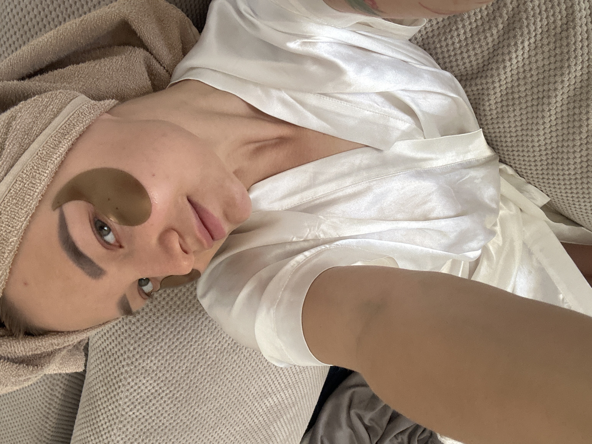 A woman is lying on a light brown textured surface, possibly a sofa or bed, wearing a white satin robe. She has a light brown towel wrapped around her head and under-eye masks applied. She is looking at the camera with a slight smile. The setting appears to be indoors. There is no information to determine the location in Budva, Montenegro.