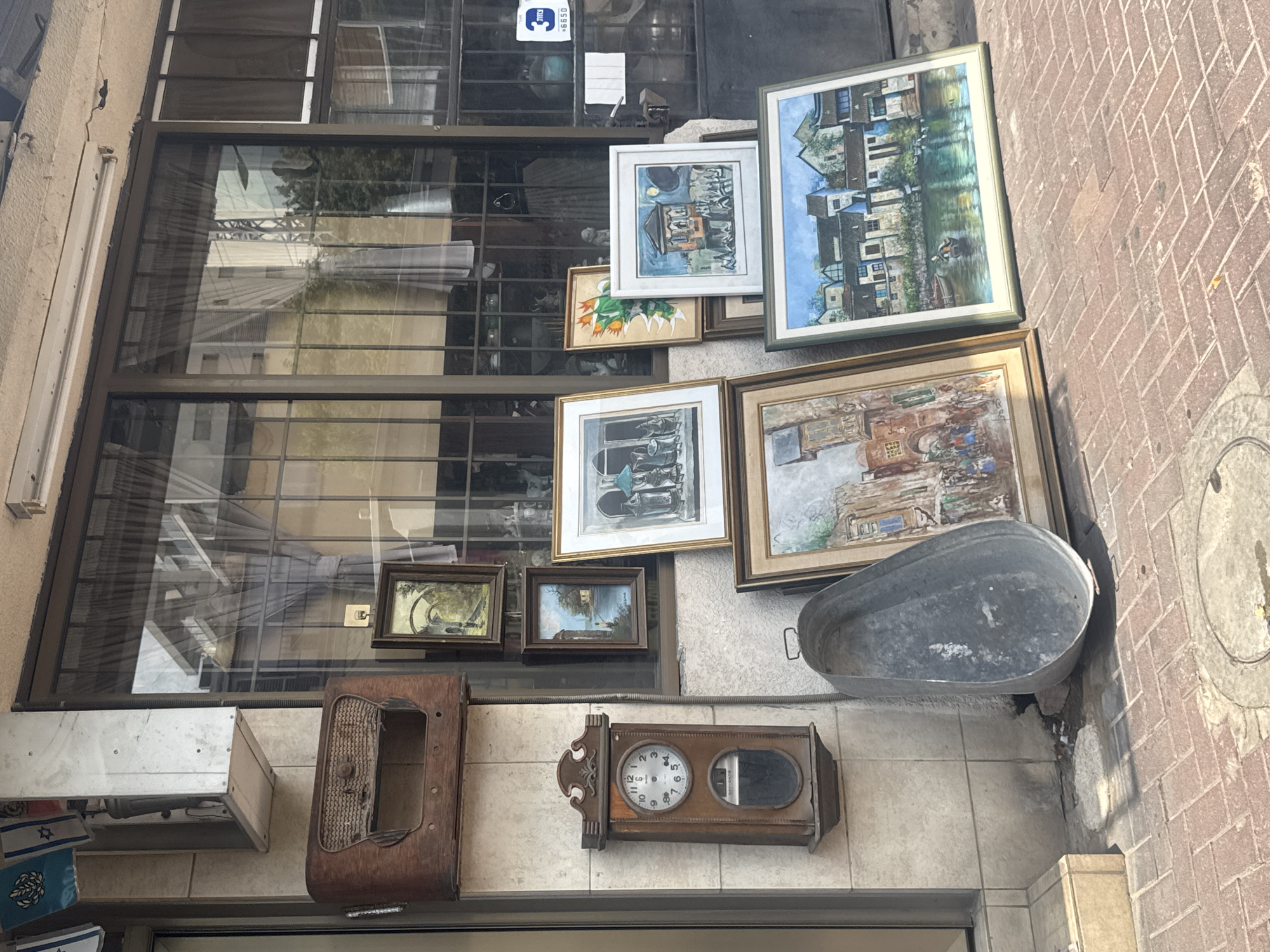 This image shows a sidewalk scene in front of a shop in Givatayim, Israel. The shop's display includes a vintage wooden radio, a pendulum clock, several framed paintings, and a large metal basin. The paintings depict various scenes, including landscapes and village life. The shop window, with its security bars, reflects buildings and trees. The sidewalk is paved with brick, and a manhole cover is visible. The overall impression is one of a quaint, somewhat cluttered, storefront offering antique or artistic items.